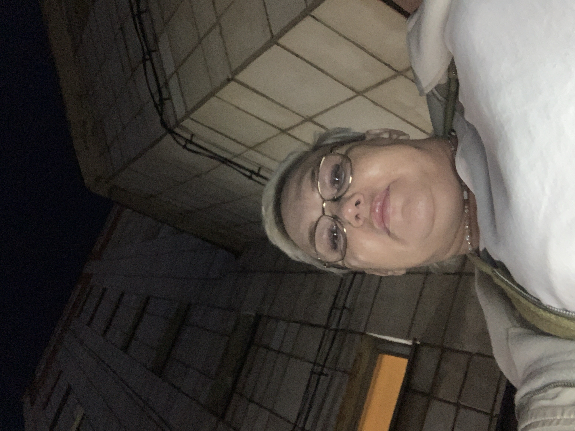 A person with gray hair and glasses is shown in a selfie-style photograph. The person is wearing a white top and jacket with a beaded necklace. The background appears to be an industrial or commercial structure. The lighting is dim. The person is oriented to the right. Based on the prompt, the location is the city of Chaikovsky, Russia, however, there is no confirmation of this in the image.