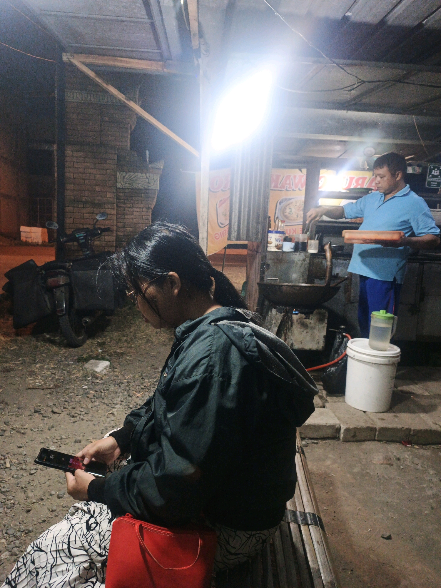 In Kintelan, Indonesia, a dimly lit outdoor eatery operates at night. A person is seated on a wooden bench, engrossed in their smartphone, with a red bag at their side. Behind them, a motorcycle is parked. In the background, a man in a blue shirt stands over a makeshift stove, attending to cooking in a wok, suggesting he is the vendor. The environment is rustic, with brick walls, wooden structures, and gravelly ground, illuminated by a bright overhead light.