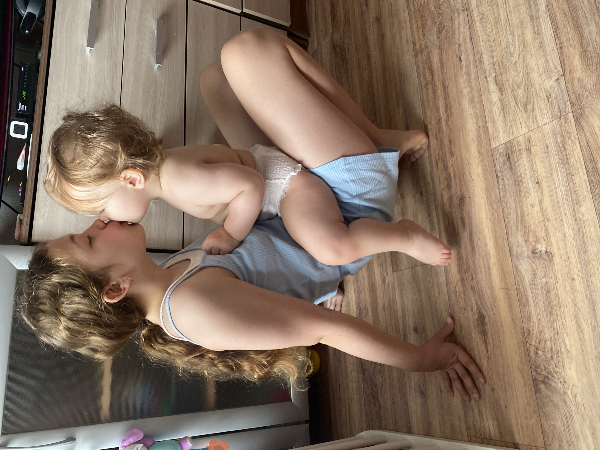 A photograph shows a woman and a toddler in an indoor setting. The woman is lying on her back on a wooden floor, wearing a light blue tank top. Her long, wavy brown hair is spread out beneath her head. The toddler, wearing a diaper and blue shorts, is lying on top of her, leaning over to kiss her on the lips. The setting appears to be a kitchen or living area, with wooden cabinets visible in the background.