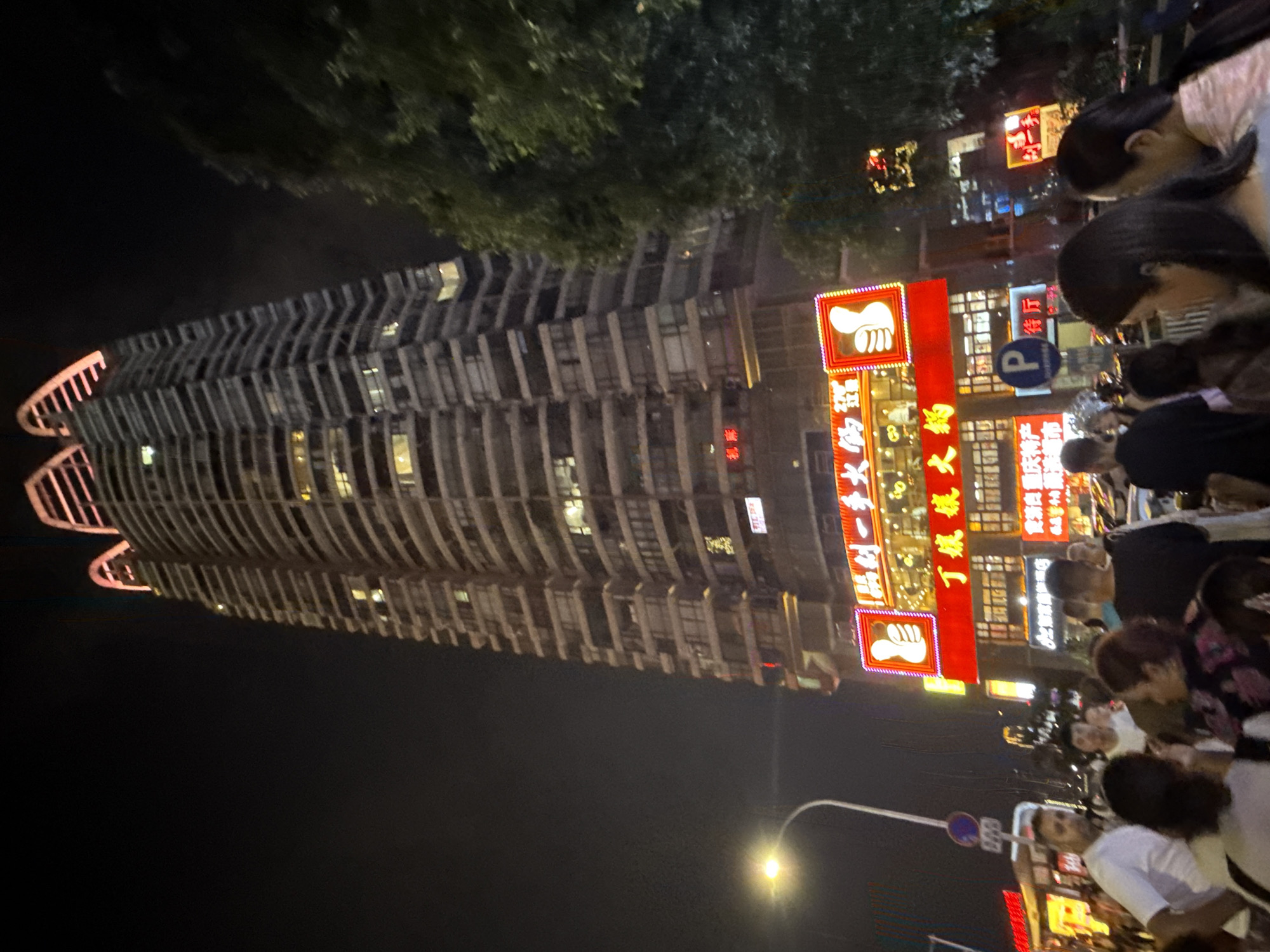 In the Yuzhong District of China, a towering skyscraper with a distinctive, illuminated crown stands prominently at night. The building features numerous floors with visible windows, some lit from within, suggesting occupancy. In the foreground, a crowd of people is gathered, indicating a public space or event. The scene is illuminated by streetlights, the building's lights, and bright neon signs adorning the lower levels of the skyscraper. One particularly large red sign features white text and a graphic of a thumbs-up symbol. Another sign, also red, displays white Chinese characters. A street lamp with a curved pole is visible to the left, and a traffic sign with a blue circle and white 'P' is present. The overall atmosphere is that of a busy urban environment at night.