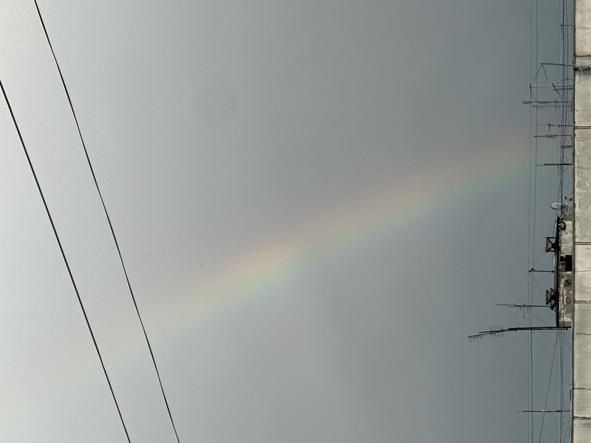 The image shows a view of the sky with a visible rainbow. Power lines are visible on the left side of the frame. On the right side, there's a concrete structure with multiple antennas attached to it. The overall environment appears overcast. The location is Unknown, Unknown.