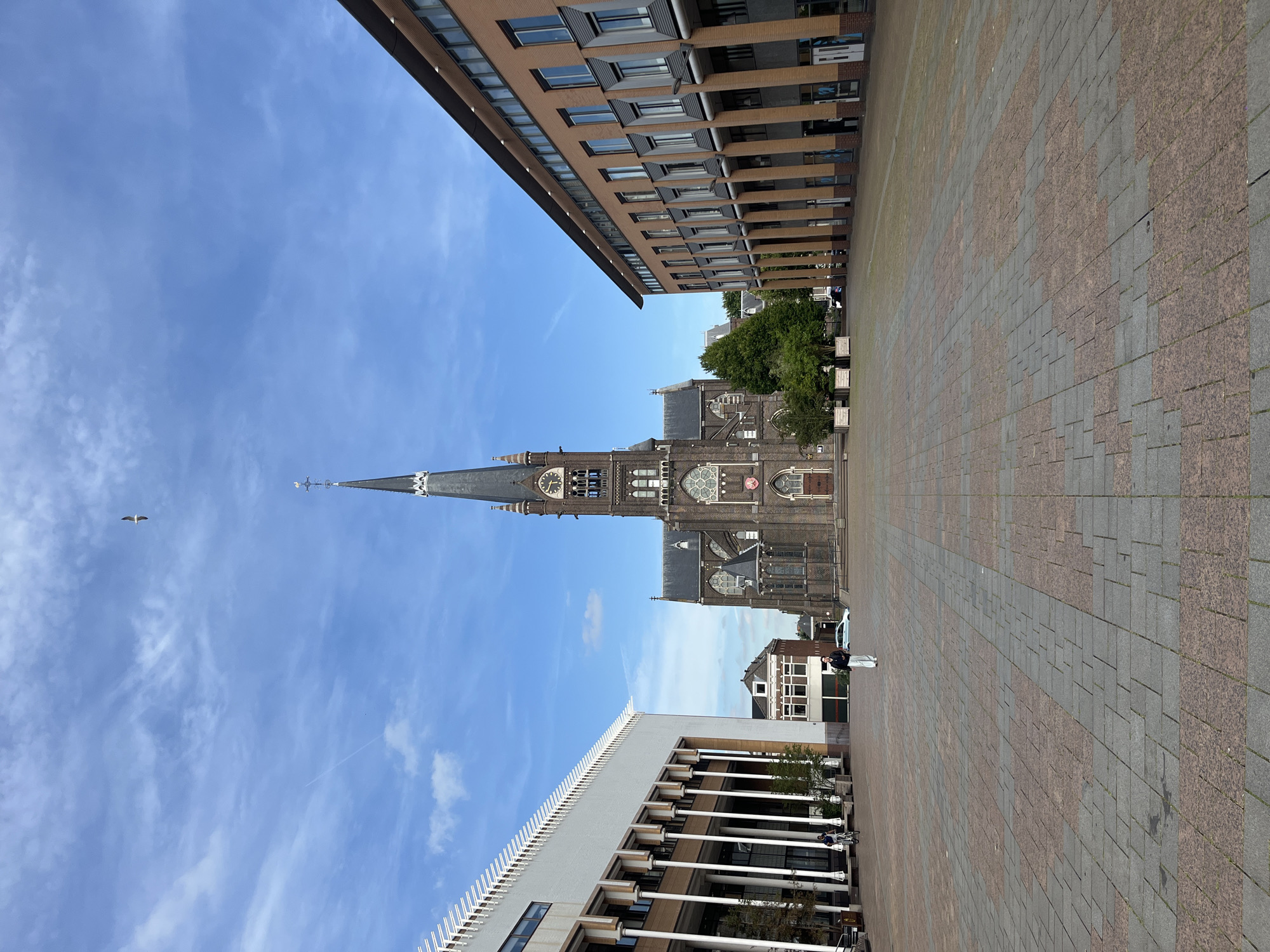 In an open, paved plaza, a large Gothic-style church with a tall spire is centrally located, facing forward. Two modern buildings flank the church, one on the left with a white facade and columns, and another on the right made of brick with numerous windows. The sky is blue with scattered clouds, and a bird is visible flying high above. A few people are present in the plaza: one person is walking their bicycle on the left side, another person is walking towards the church in the center, and a white car is parked in the background. The plaza is paved with a pattern of reddish-brown and gray square tiles.