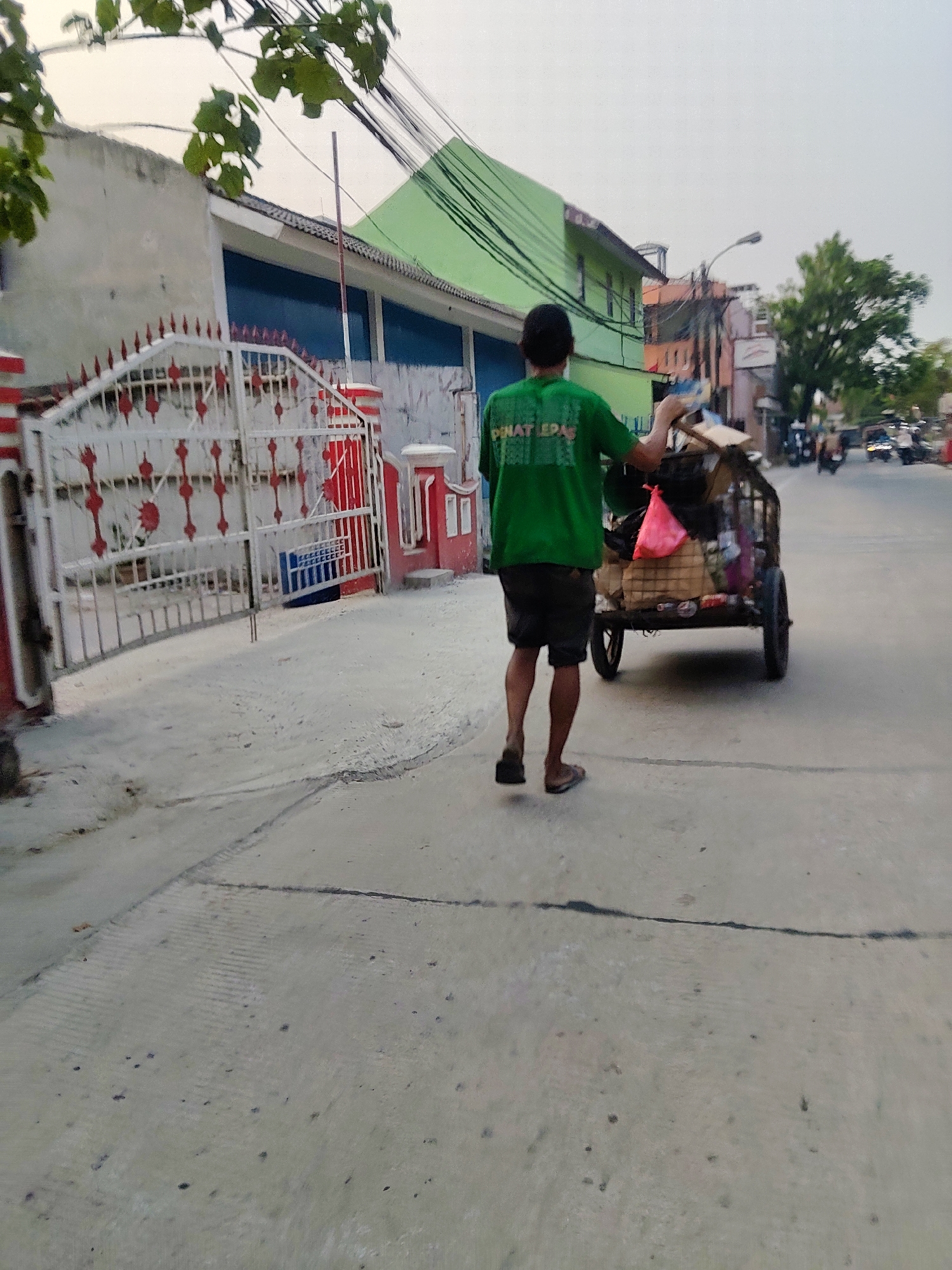 In Mekarmukti, Indonesia, a person wearing a green t-shirt and dark shorts walks on a paved street, pulling a two-wheeled cart filled with various items. The cart is laden with bags, cardboard boxes, and what appears to be miscellaneous debris. The person is seen from behind, with their left hand on the cart's handle. The street is lined with buildings, including one with a green facade and another with a white and red striped gate. Overhead, electrical wires crisscross the sky, and some leafy branches extend from the upper left of the frame. In the background, other vehicles, including motorcycles and a small car, are visible on the street, suggesting a moderately active urban environment. The overall lighting suggests it is daytime, possibly late afternoon given the ambient light.