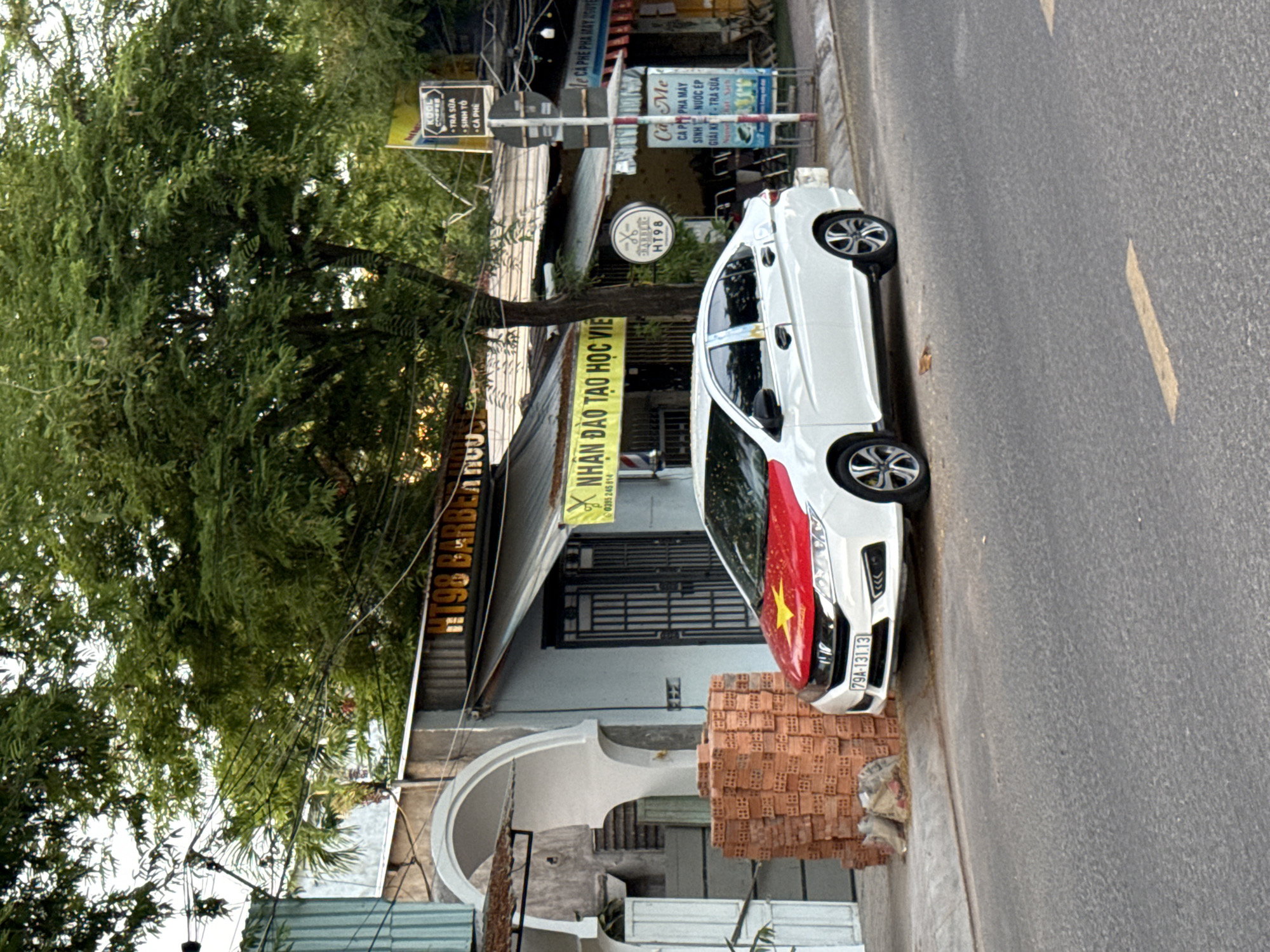 The image depicts a street scene in Nha Trang, Vietnam. In the foreground, a white sedan is parked on the side of the road. The car has the Vietnamese flag, a red flag with a yellow star, displayed on its hood. To the left of the car, a pile of red bricks is stacked on the sidewalk. Behind the car, a building with a storefront is visible. A yellow banner with Vietnamese text is hanging above the storefront. Several trees with dense green foliage line the street. The overall environment suggests a commercial or residential area in a Vietnamese city.