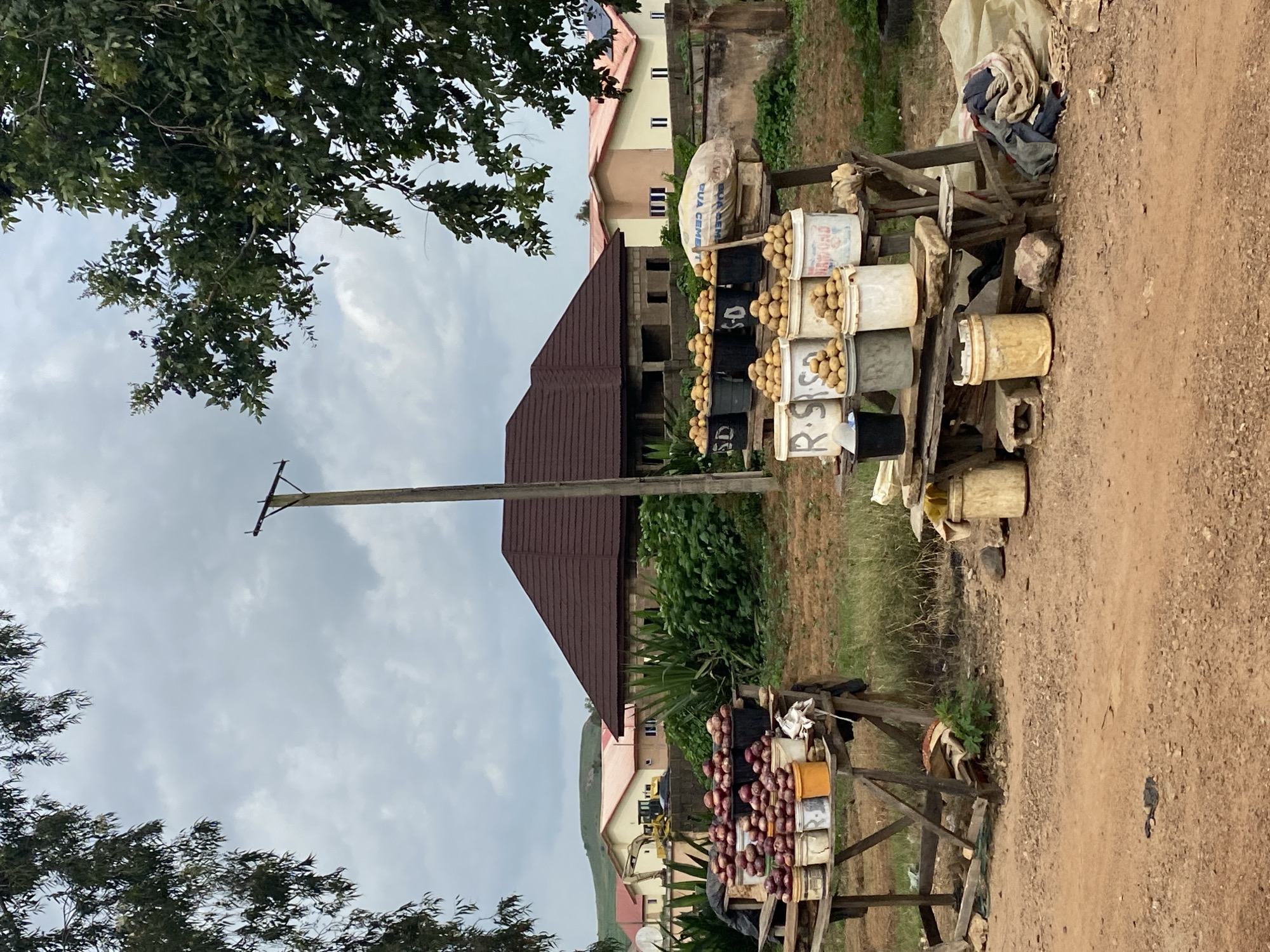 This is a medium-shot, eye-level, outdoor photograph taken on a cloudy day in the city of Riyom, Nigeria. The image depicts a roadside fruit stand with various produce displayed in buckets and on wooden tables. In the foreground, there is a dirt road.
To the left, a wooden stand holds multiple buckets filled with what appear to be red onions. To the right, another wooden stand displays several buckets filled with potatoes. A large, beige bag labeled "DUA CEMENT" is also visible on this stand. Several empty buckets and some fabric items are scattered around the base of the stands.
In the background, there are several houses with red-tiled roofs. Behind the houses, rolling green hills are visible. A tall utility pole stands centrally in the mid-ground. Lush green vegetation and trees frame the scene on both sides and in the background. A yellow excavator is parked in front of one of the houses. The overall setting suggests a rural or semi-urban area.