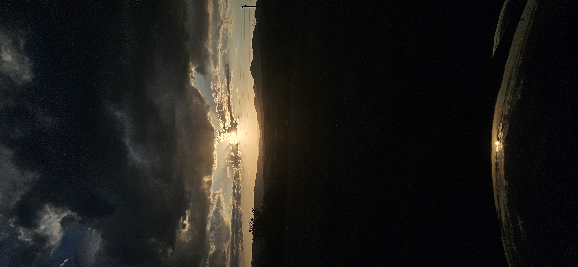 The image shows a landscape at sunset near Momin Prohod, Bulgaria. Dominant dark storm clouds fill the left side of the frame, transitioning to lighter, sunlit clouds near the center. The sun is partially obscured by the clouds, creating a glowing effect with visible sun rays. A mountain range is visible on the horizon in the middle of the frame. Trees are silhouetted against the fading light. Power lines are visible in the sky. A dark, undefined area occupies the right side of the image.