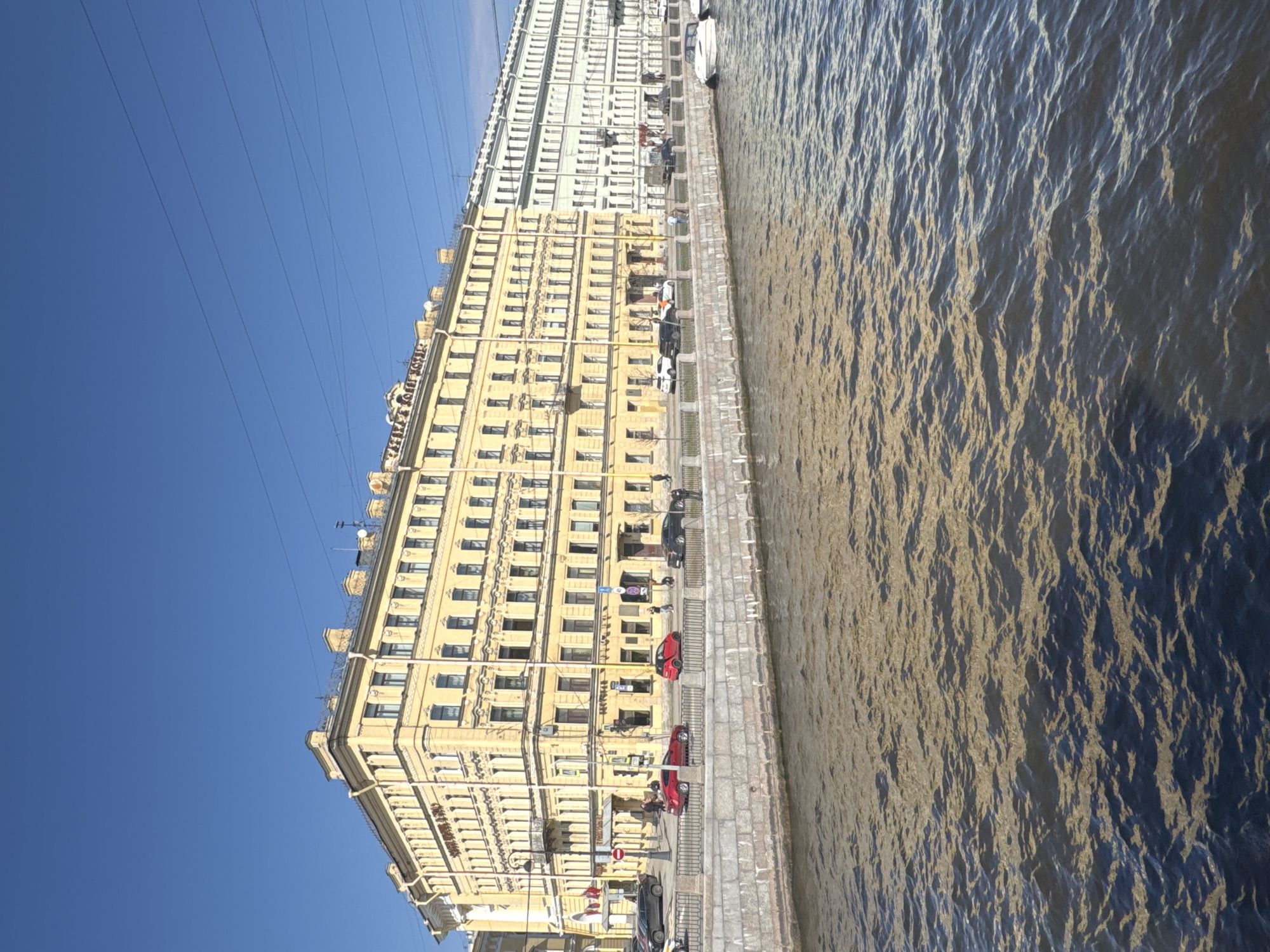 The image depicts a view of a building along the embankment of a river in Saint Petersburg, Russia. The building is a tall, yellow, ornate structure with numerous windows. Adjacent to this building is a similar, though slightly lighter-colored, building. A paved walkway runs along the edge of the embankment, with parked cars and pedestrians visible. The river appears choppy, with the sunlight reflecting off the surface. The sky is clear and blue. The scene is bathed in daylight, with the sun casting shadows on the buildings and the river. Power lines run across the sky.