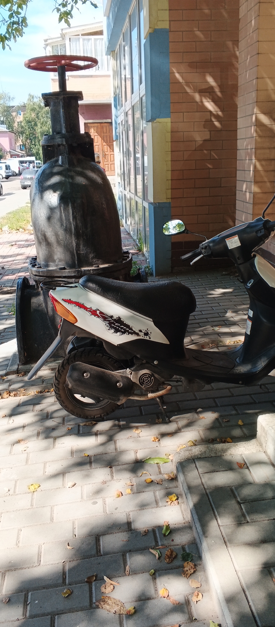 In Smila, Ukraine, a black and white scooter is parked on a sidewalk next to a large, industrial pipe with a red wheel on top. The scooter has a white panel with red and black graphics. The sidewalk is paved with gray bricks and scattered with fallen leaves. In the background, a building with large windows and brick walls is visible. The sky is blue and partly cloudy.