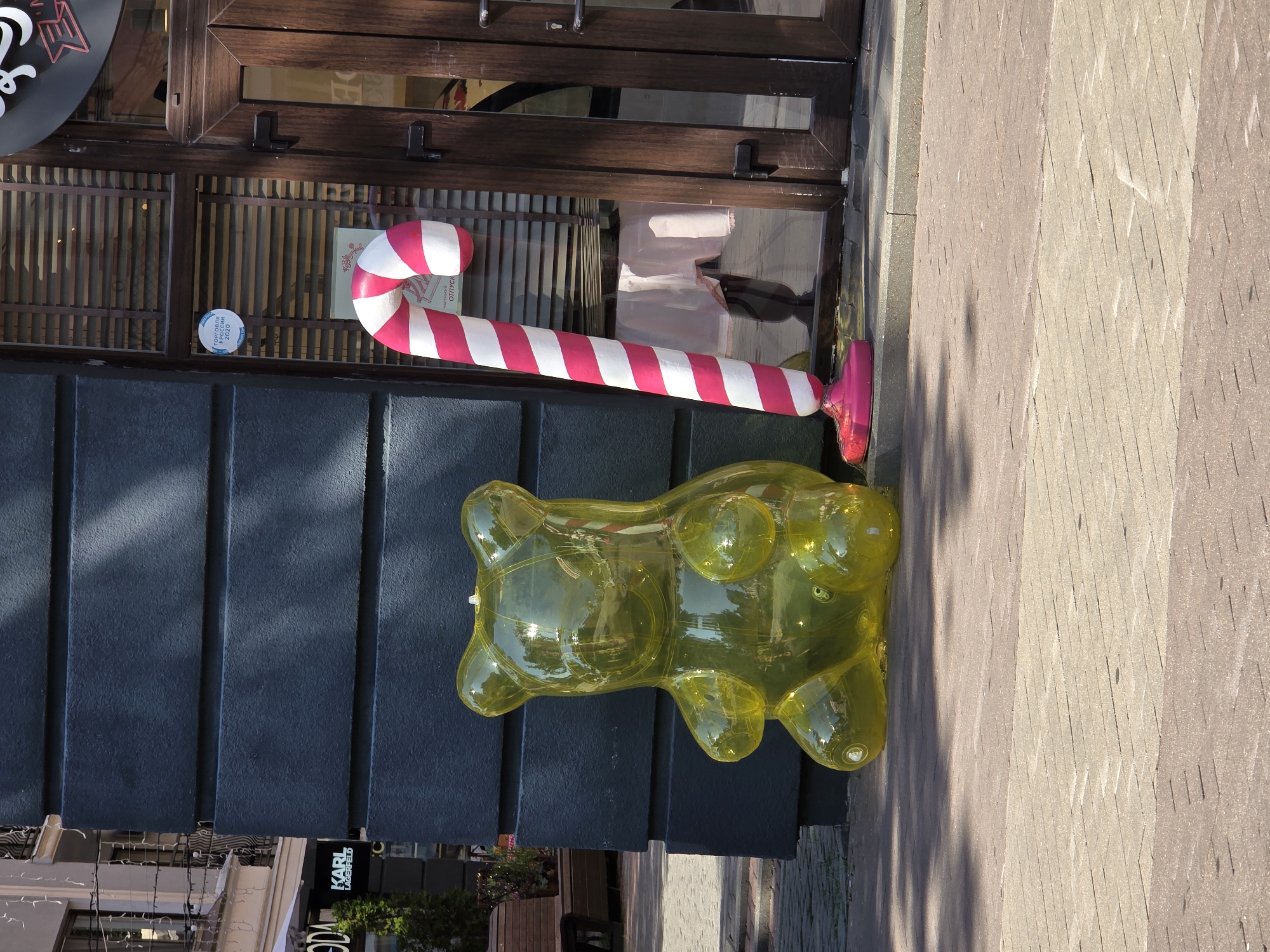 A yellow, inflatable gummy bear figure sits on a paved sidewalk in front of a dark gray building. To the right of the gummy bear is a large, striped candy cane decoration with pink and white stripes, standing upright and leaning slightly. The candy cane has a bright pink base. Behind the decorations is a building entrance with dark wooden doors and a large window covered by horizontal blinds. A small, round sticker is visible on the window. Sunlight casts shadows from nearby trees onto the ground and the building. In the background, a portion of another building with a light-colored facade and balconies can be seen. The overall environment appears to be a commercial street.