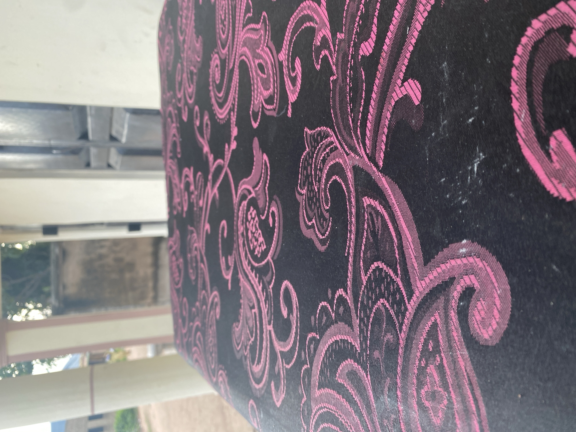The image is a close-up, angled shot of a black fabric with a pink paisley pattern. The fabric appears to be part of an upholstered object, possibly furniture, given its texture and the way it is draped. The paisley design is intricate and consists of swirling, leaf-like shapes. The pink color of the pattern stands out against the dark background.
In the background, out of focus, there are hints of an outdoor setting. A light-colored structure, possibly a wall or building, is visible, with some greenery and possibly other buildings in the distance. The lighting suggests it is daytime, with natural light illuminating the scene. The overall impression is one of everyday life, with the focus sharply on the textured fabric. There is no visible text in the image. The location context of Jalingo, Nigeria, does not provide specific visual cues within the image itself, but it grounds the observation in a particular geographic setting.