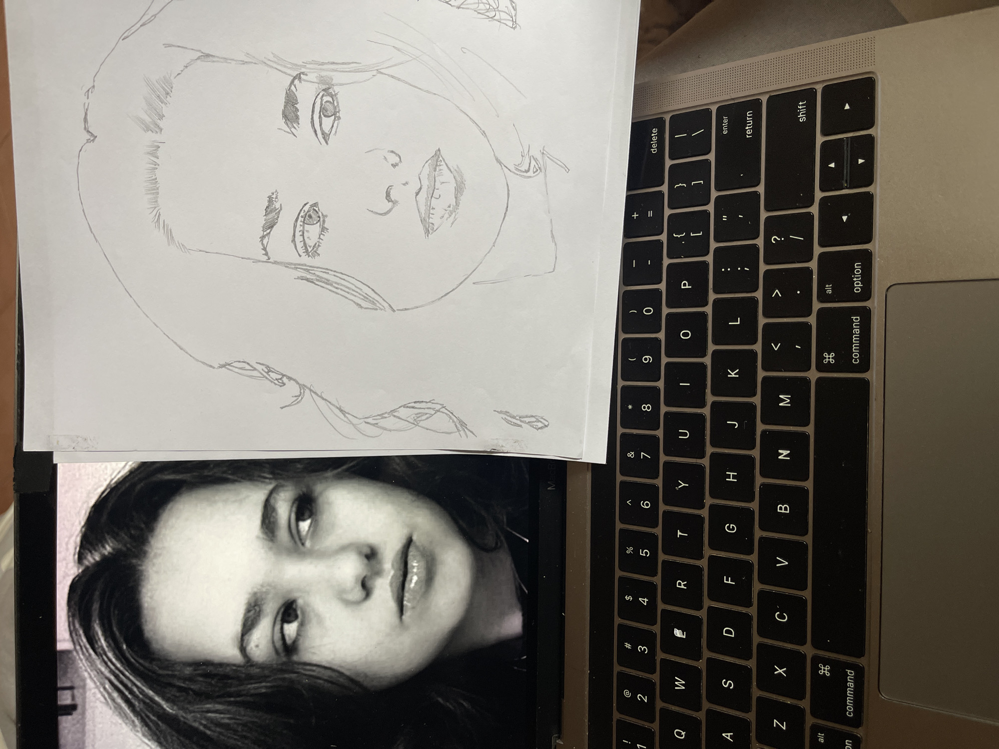 A photograph depicts a pencil drawing of a person's face laid on top of a laptop. The laptop keyboard is visible, occupying the right side of the frame, with keys and the trackpad clearly seen. The drawing is on a white piece of paper, positioned over the laptop screen which displays a grayscale photograph of a person's face. The drawing appears to be an attempt to replicate the image on the screen. There is no indication in the image of the city of Jos, Nigeria.