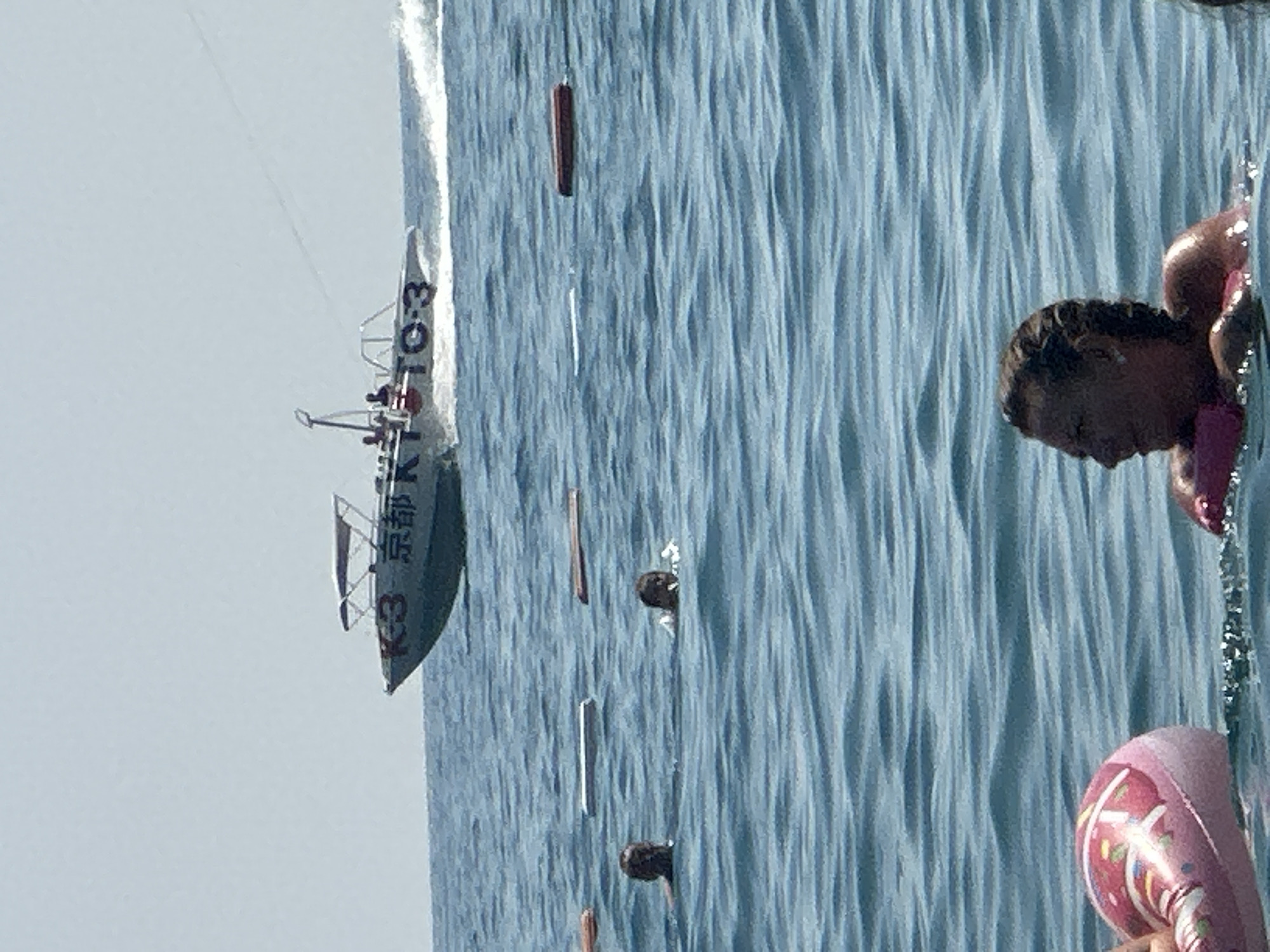 A motorboat, marked with "K3 京都 K-3", speeds across the blue, choppy water of the sea, leaving a white wake. The boat carries several passengers. In the foreground, two individuals swim, their heads visible above the water. In the lower left corner, a pink inflatable ring with a pattern is partially submerged. The scene appears to be set on a sunny day with a clear, pale blue sky. The location is the sea off the coast of Boreti, Montenegro.
