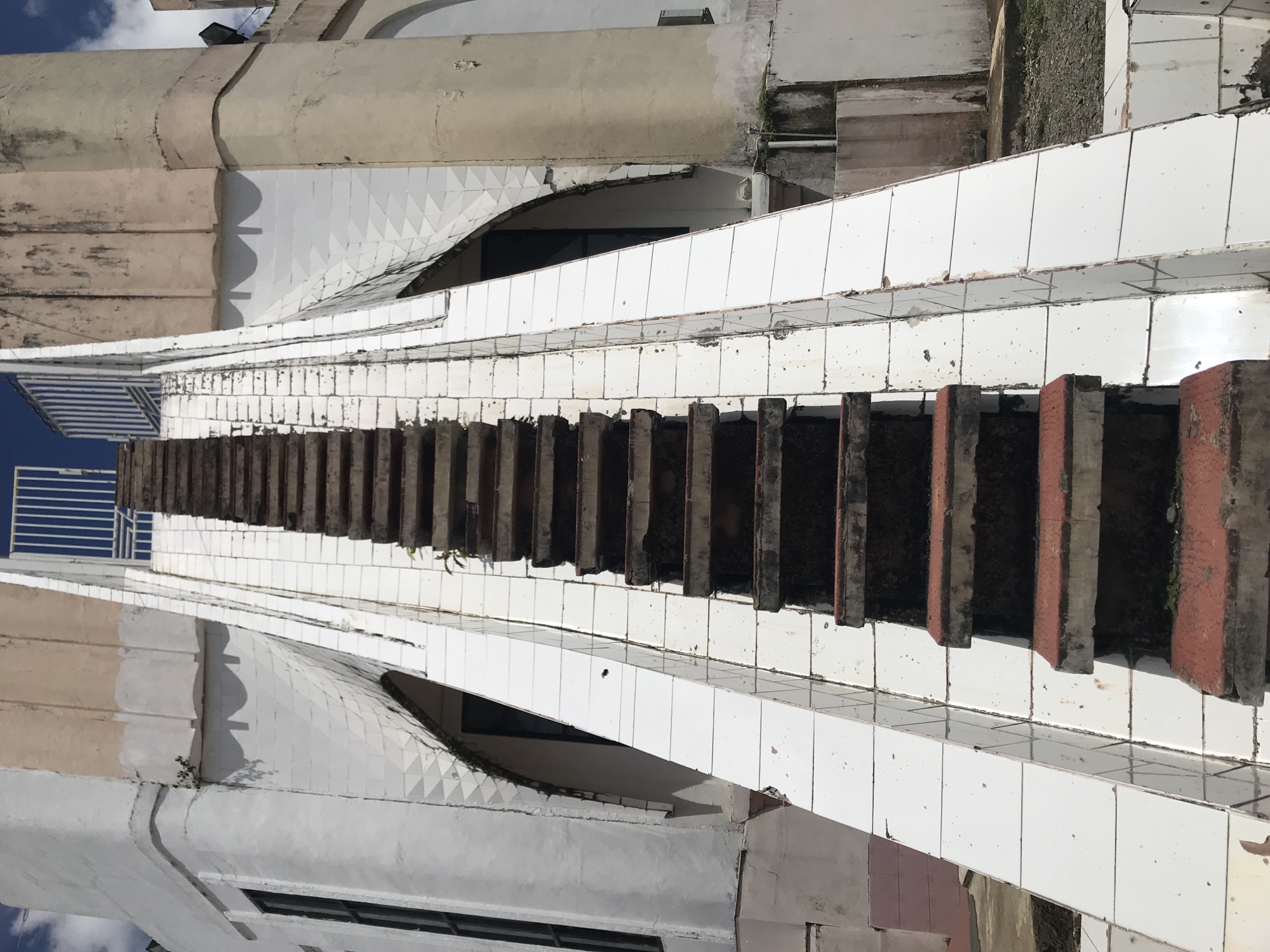 An upward-angled shot depicts the side of a building in Jantho, Indonesia. A narrow staircase lined with white tiles is prominently featured, its steps alternating between dark grey/black and some with red-brick colored edges. A metal railing is visible at the top of the stairs. The building itself is a mix of curved and angular structures, with white triangular tiling on a curved surface and other sections with smooth, white surfaces. The building's overall condition suggests some wear and tear. The sky is partially visible in the upper left, indicating an outdoor setting.