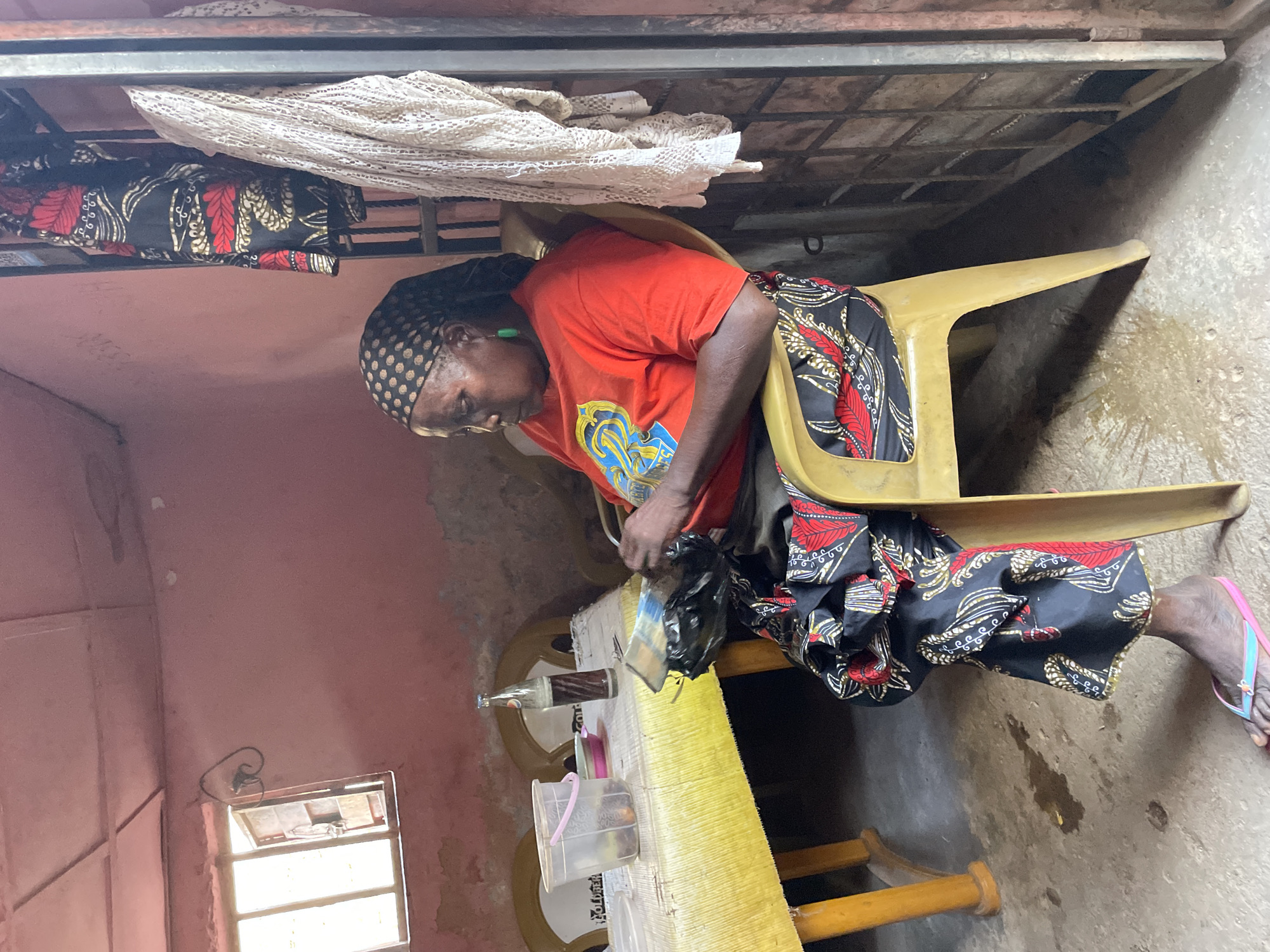 An elderly woman in Jos, Nigeria, wearing a patterned headscarf, an orange t-shirt, and a black skirt with red and gold designs, is sitting on a yellow plastic chair in an interior setting. She is leaning forward, seemingly handling a black plastic bag. A long, yellow table is in front of her, holding various items, including a bottle, a container, and other objects. The room has pink walls, and a small window provides light. A metal bunk bed frame is visible above her, with a white crocheted cloth and another patterned garment hanging on it. The floor appears to be concrete. The overall environment looks somewhat worn and simple. The woman is wearing pink and blue flip-flops.