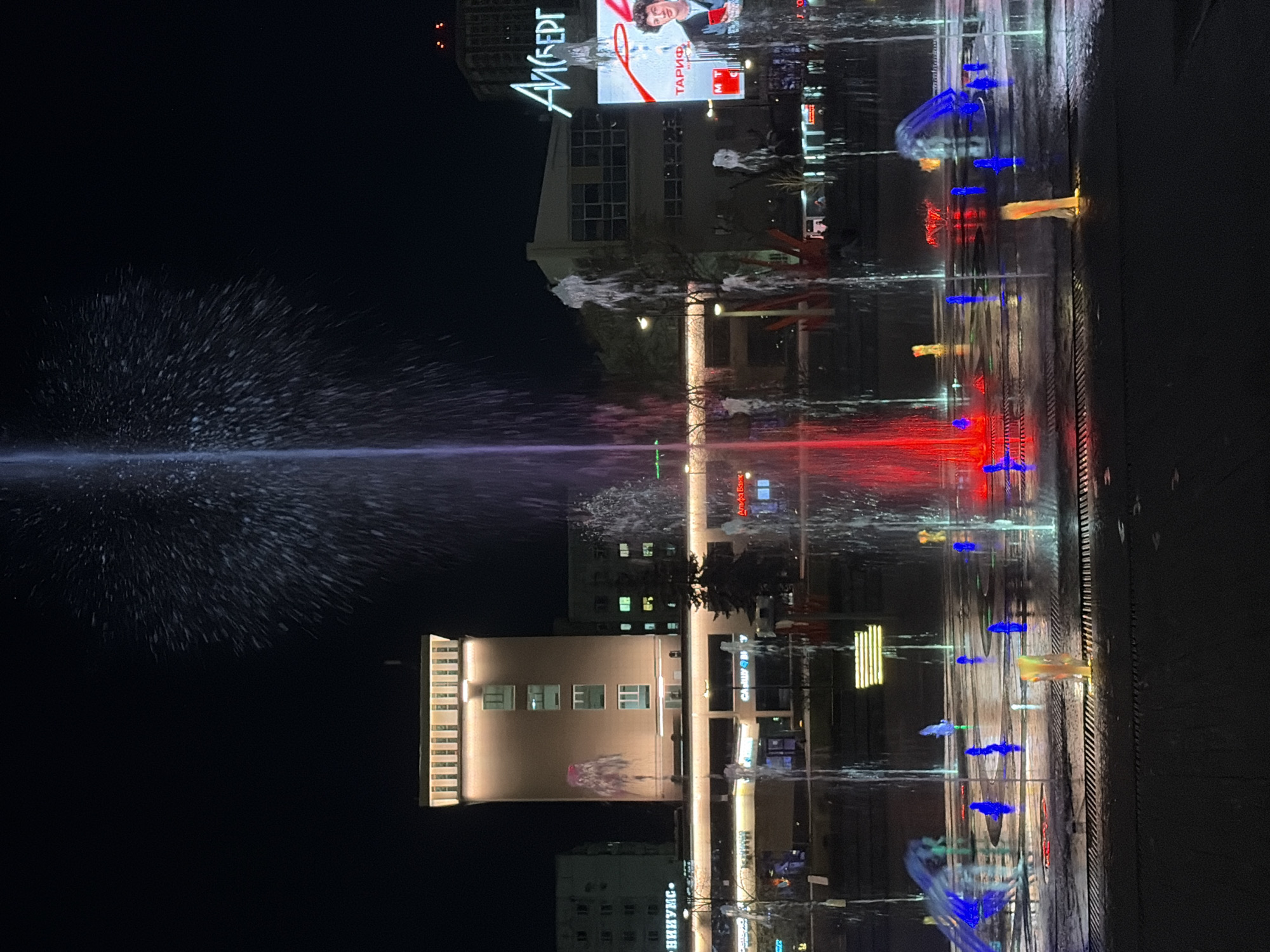 At night, the photo captures a water fountain illuminated with purple and red lights in Perm, Russia. The fountain's spray pattern is widespread and large. In the background are buildings and a billboard advertising "Aйсберг" and "MTC Тарифы." Other lights illuminate the structures, creating reflections on the wet ground around the fountain. The scene is urban, with the focus primarily on the fountain and its surrounding urban environment. The image is horizontally oriented.