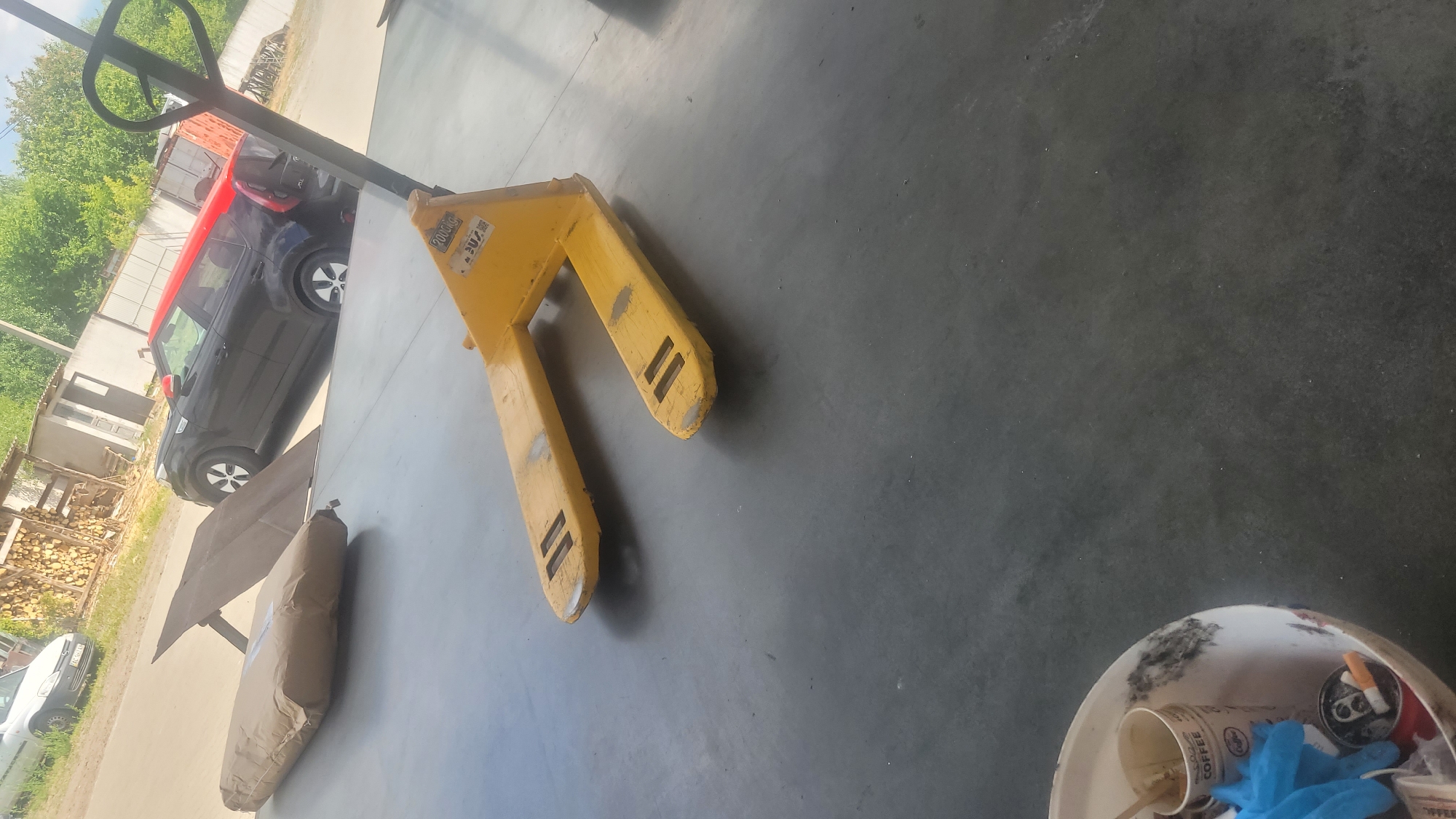 An outdoor scene shows a yellow pallet jack on a dark gray concrete surface. In the background, a black car with a red roof is parked. To the left of the car, a stack of firewood is visible next to a building. Further left, a white car is parked. In the lower left corner of the image, a white bucket contains a disposable coffee cup, blue gloves, and cigarette butts.