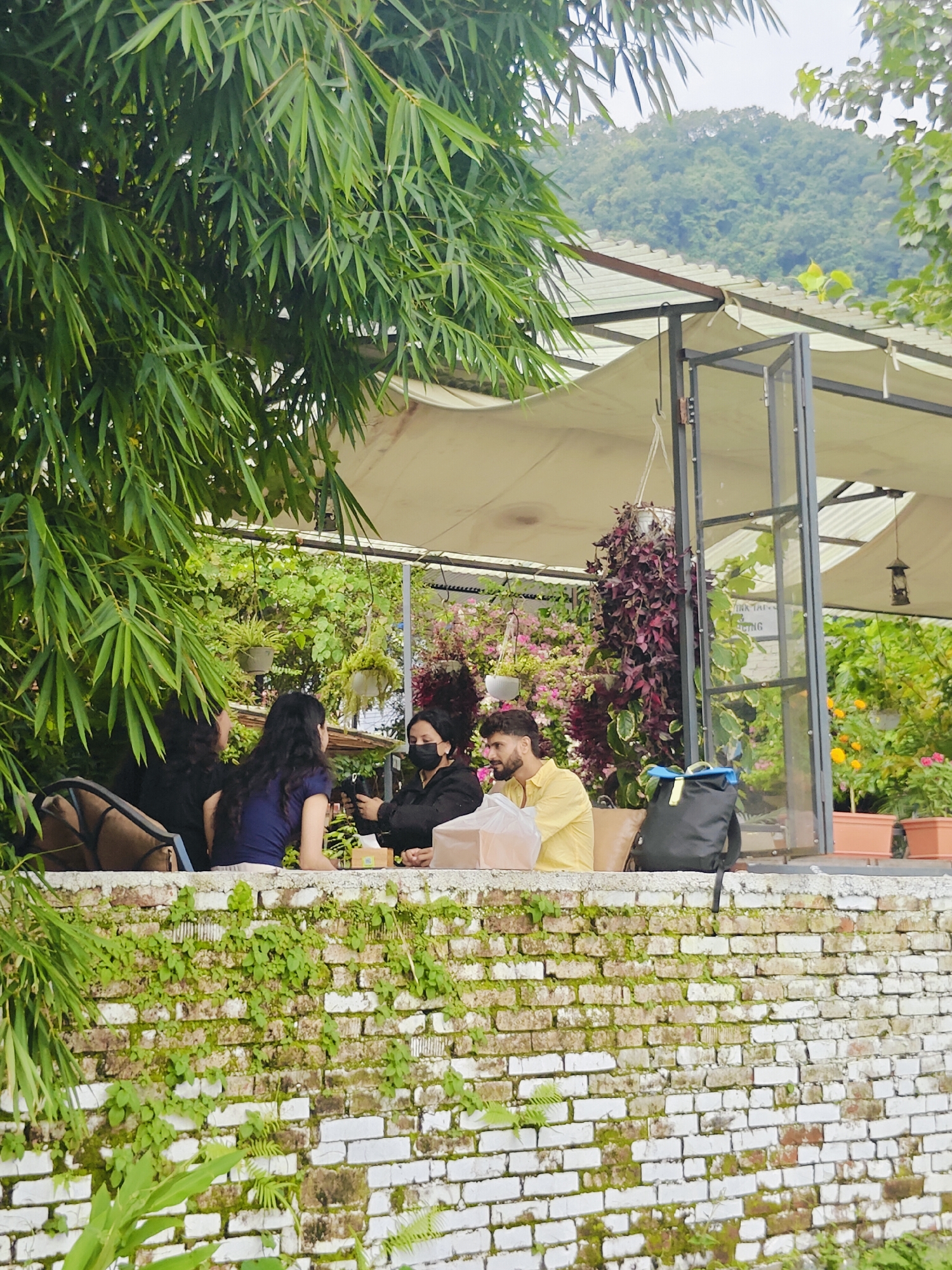 The image depicts a group of people seated at a table on a patio overlooking a lush, green hillside, likely in Pokhara, Nepal. The foreground features a weathered brick wall covered in moss and vines. Behind the wall, four individuals are visible, three women and one man, seated around a table. The woman on the far left is mostly obscured by foliage and a chair. To her right, a woman with long dark hair is seated, wearing a dark top. Next to her, a woman wearing a blue t-shirt is facing away from the camera. The woman closest to the viewer is wearing a black mask and a dark jacket, holding a phone. The man, wearing a yellow shirt, is seated to her right, looking towards the other individuals. A black backpack with blue straps rests beside him. The background reveals a covered outdoor seating area with a thatched roof. Beyond the seating area, dense vegetation and hanging plants are visible, and further in the distance, a heavily forested mountainside under an overcast sky. The overall impression is a tranquil, natural setting.