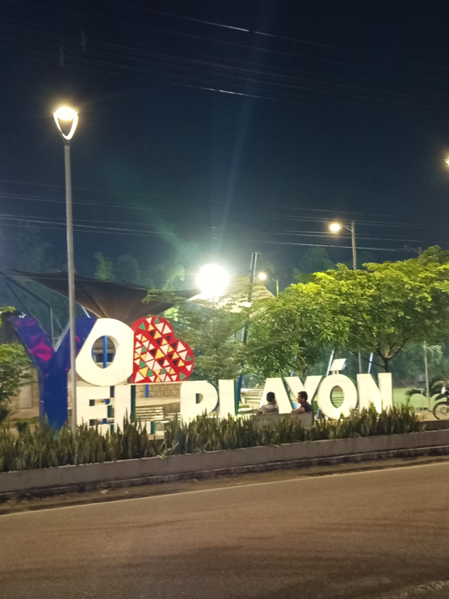 This is a nighttime, eye-level, medium shot of the "YO EL PLAYÓN" sign in El Playón, Santander, Colombia. The sign is illuminated by streetlights and is positioned in front of lush green trees. The large, white letters spelling out "EL PLAYÓN" are prominent in the foreground, with a stylized red, white, and yellow heart icon integrated into the design. The letters "YO" are partially visible on the left side of the frame, with the "Y" being blue and the "O" being white and circular. Two individuals, a male and a female, are seated on a bench behind the sign, facing towards the right. A motorcycle is parked to the far right of the frame. The background consists of dark trees and the night sky with visible power lines and streetlights. The scene appears to be a public park or recreational area within the city.