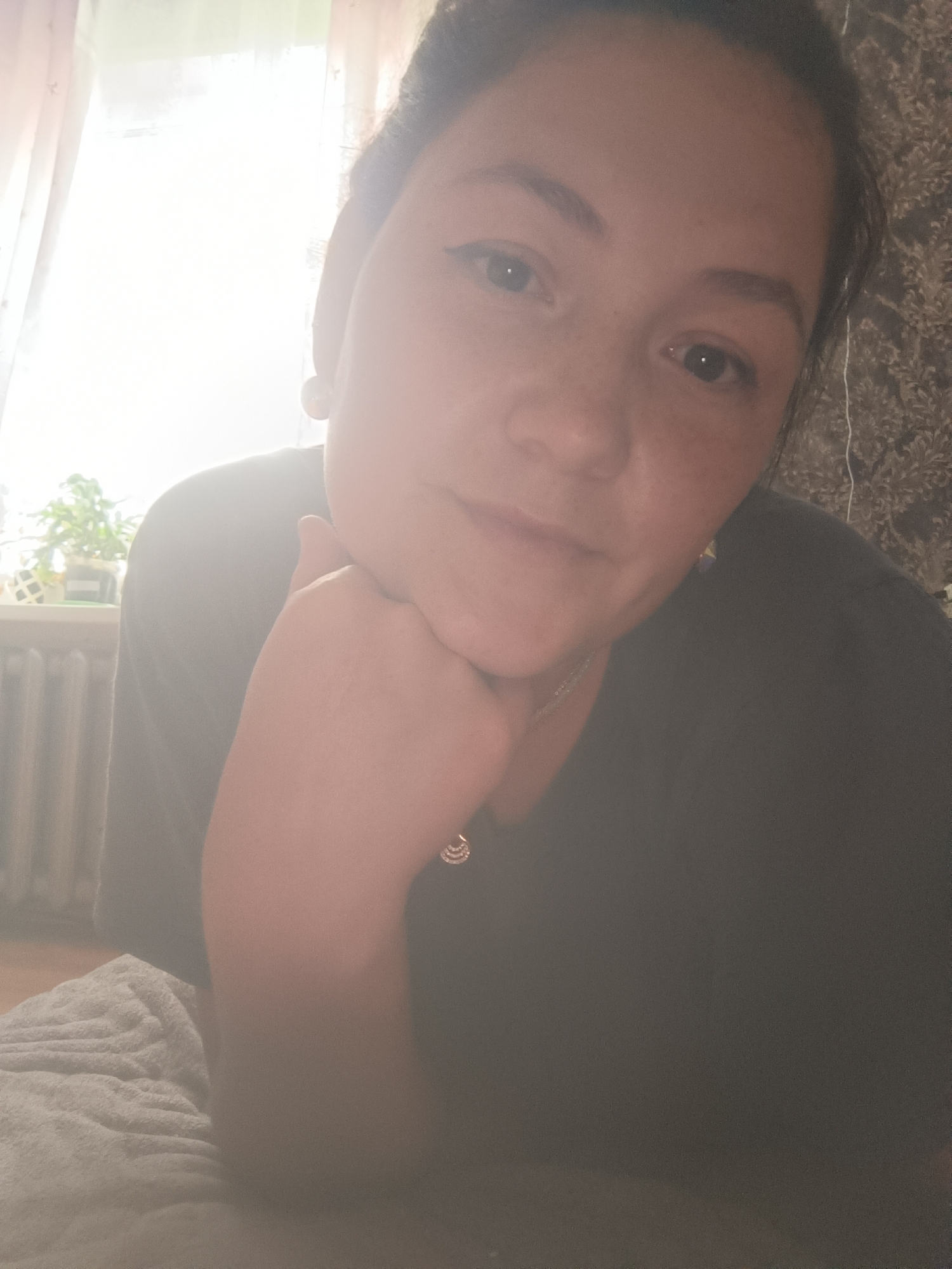 The image is a close-up, indoor selfie of a woman with her head resting on her hand. She has dark hair pulled back, and she is wearing a dark-colored shirt. She has earrings and a necklace on. In the background, there is a window with sheer curtains and a radiator, suggesting an apartment or house. To the right of the woman, a patterned wallpaper is visible. The lighting suggests it is daytime, with light coming in from the window. The overall scene appears to be a casual, personal moment captured in what is likely a residential setting within the city of Karsha, Russia.