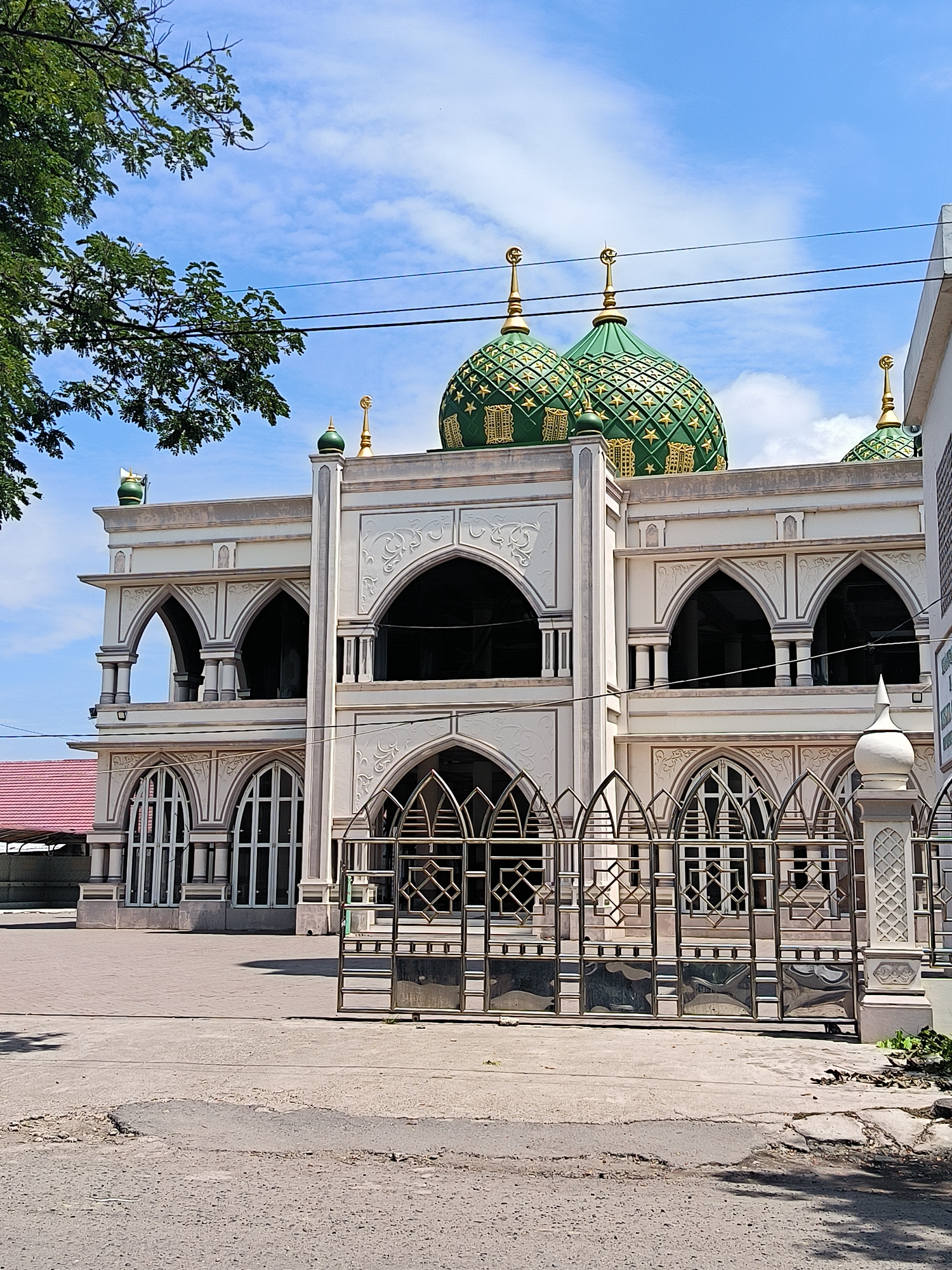The image depicts the exterior of the Baiturrahman Grand Mosque in Lhokseumawe, Indonesia. The mosque features a prominent white facade adorned with intricate architectural details, including arched windows and decorative patterns. Two large, green domes, each topped with a golden crescent moon, dominate the rooftop. A smaller dome is visible on the right side of the structure. The mosque is surrounded by a paved courtyard and a decorative metal gate. Overhead, power lines stretch across a bright blue sky dotted with fluffy white clouds. A leafy tree branch extends into the frame from the upper left, casting a shadow on the scene. The ground in the foreground is paved and shows some signs of wear, with a patch of asphalt appearing damaged. No people are visible in the image.
