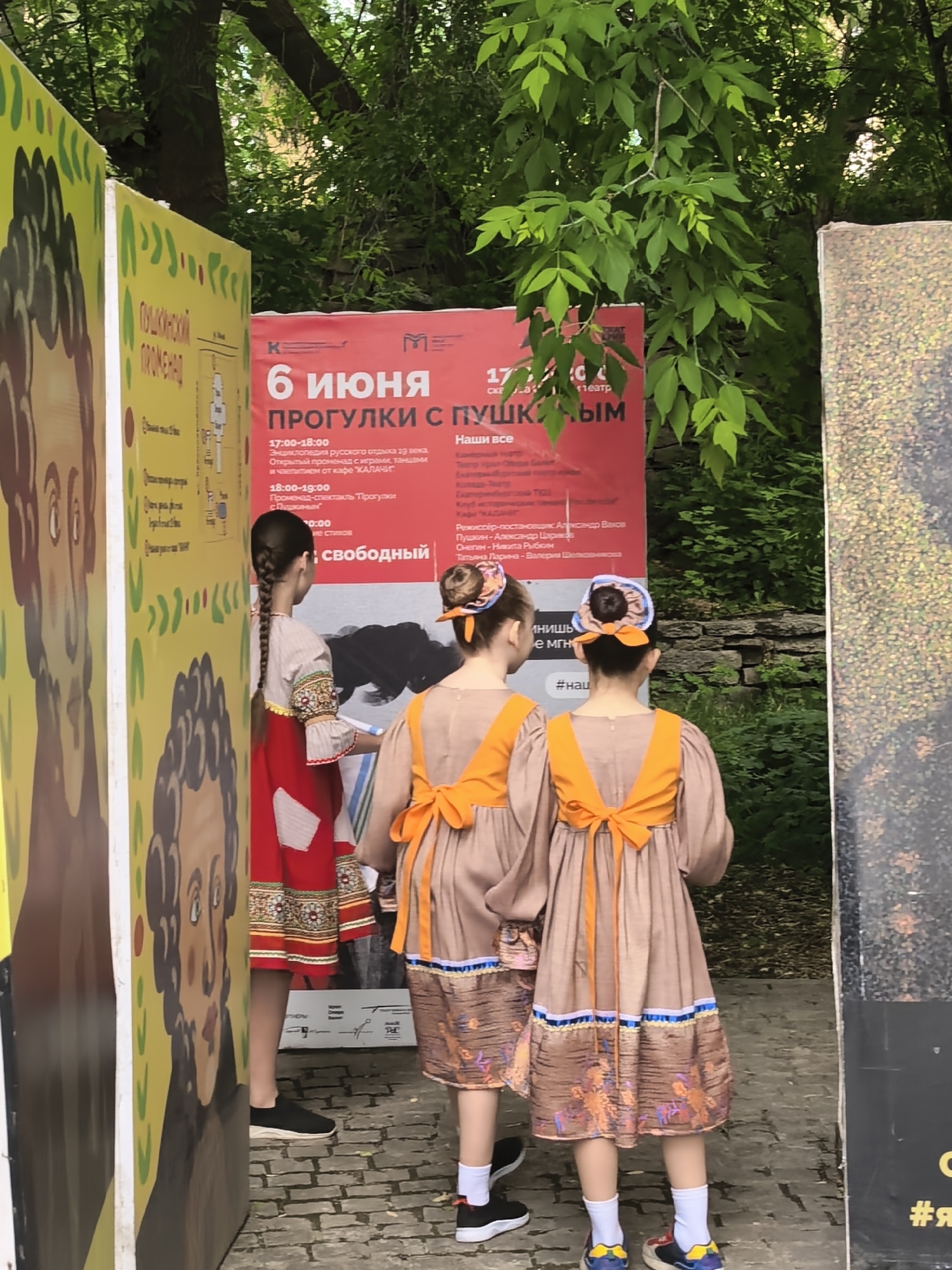 Here is a detailed description of the image:
Three young girls are standing in front of an event poster in an outdoor setting, presumably in Unknown, Unknown. The poster advertises an event called "Walks with Pushkin" on June 6th, with scheduled activities including historical reenactments, dancing, refreshments, and poetry readings.
The girls are dressed in traditional Russian clothing. The girl on the left has a long braid and wears a red and white dress with ornate trim. The two girls on the right have matching brown dresses with orange accents and bows in the back. Each wears a decorative headpiece with matching colors.
The environment includes trees in the background, suggesting the event is being held in a park or similar outdoor space. There are also other posters adjacent to the main one, but their content is partially obscured. The ground appears to be paved with cobblestones or bricks.