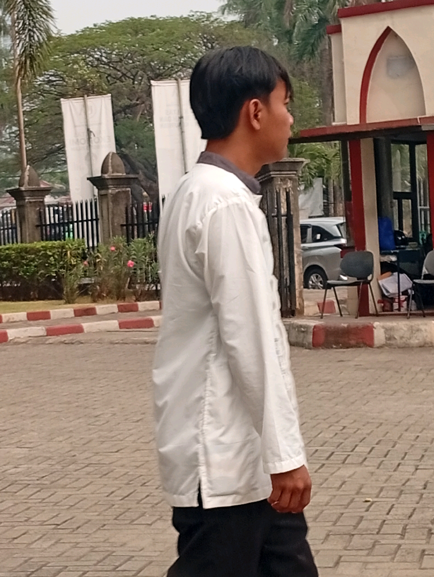 The image shows a young man, appearing to be of Asian descent, walking outdoors in a paved area. He is wearing a white long-sleeved shirt and dark pants. His hair is short and dark. In the background, there is a building with an arched entrance and red and white trim, as well as some greenery, including trees and bushes. There is also a black chair visible. The man is in profile, looking to his right. The scene appears to be in an urban or semi-urban environment. Based on the provided information, the location is identified as Kelapa Dua, Indonesia.