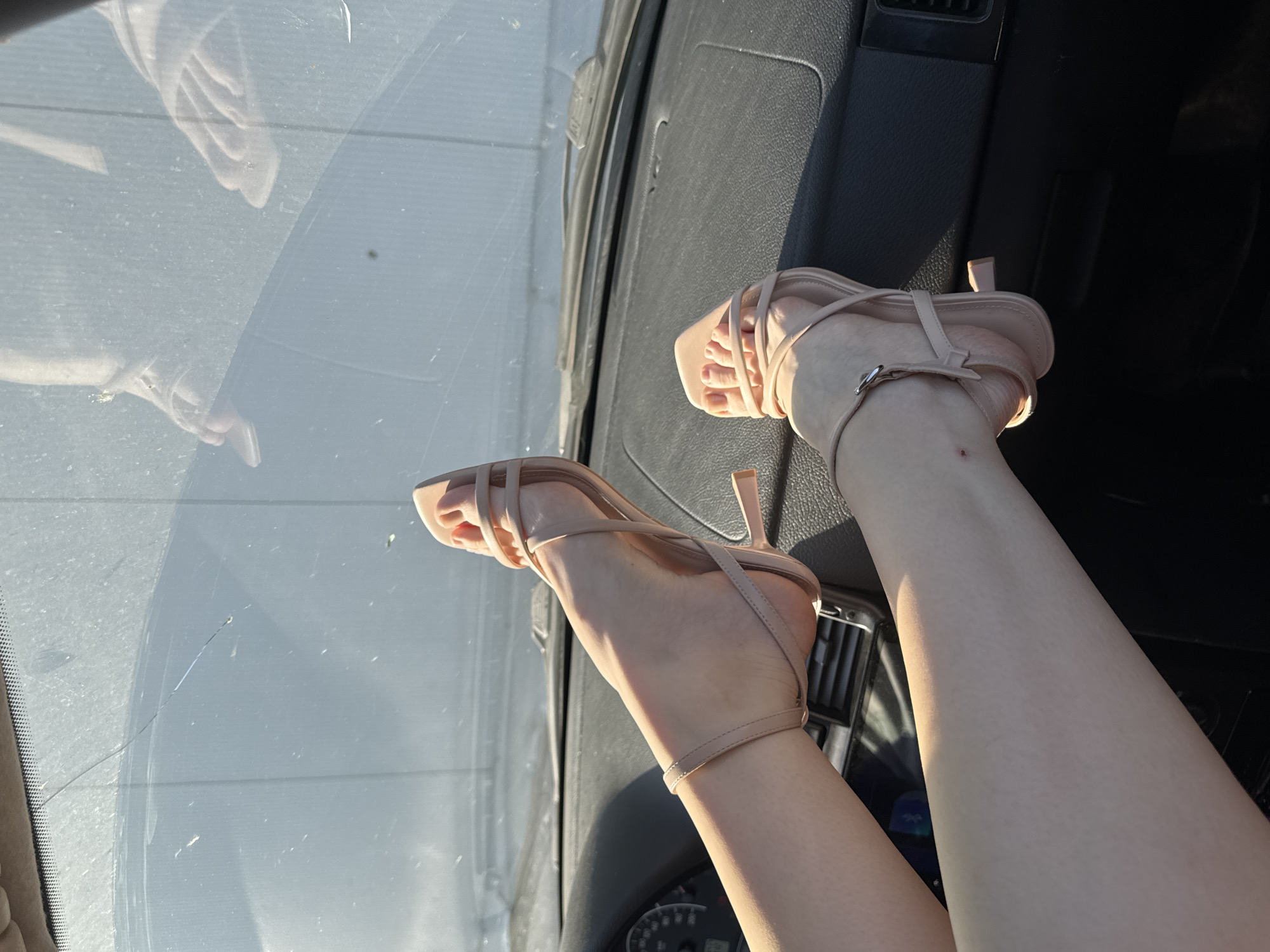 The image shows a pair of bare legs with pale skin, positioned inside a vehicle. The feet are adorned with nude-colored strappy high-heeled sandals. The legs are resting against the dark gray dashboard of the car. The background is blurred but appears to show the interior of the car and, through the windshield, a view that suggests an outdoor setting. The reflection of a figure is visible in the windshield. There is no definitive information to place this image in Perm, Russia.