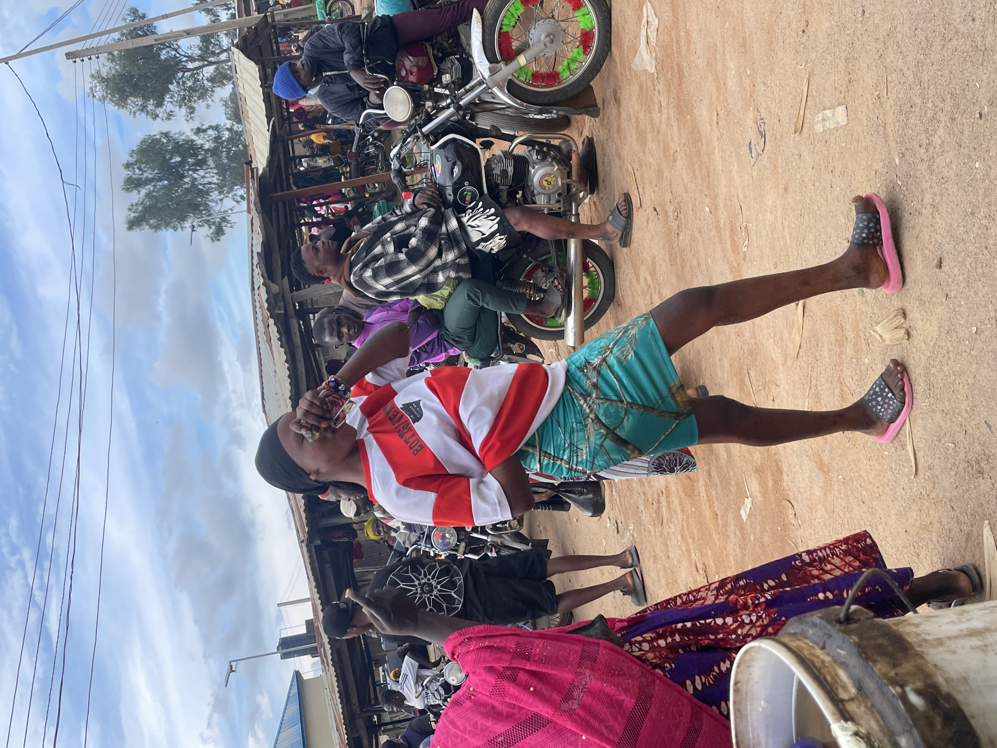 In the city of Vom, Nigeria, a young female is captured mid-action, holding a clear plastic cup to her lips as she drinks. She is wearing a red and white striped t-shirt with the text "BOTTSJEKYN" on the chest, paired with a teal-colored skirt adorned with a camouflage-like pattern of branches. Her feet are clad in pink and black slide sandals. The background reveals a bustling market scene with numerous individuals and motorcycles. A man in a black and white plaid shirt and dark shorts sits on a black motorcycle, while another man in a blue beanie and jacket is visible to the right. Stalls with corrugated metal roofs line the area, and the ground is composed of reddish-brown dirt. Overhead, a cloudy sky with power lines stretches across the frame. The image depicts a vibrant, everyday moment in a Nigerian marketplace.