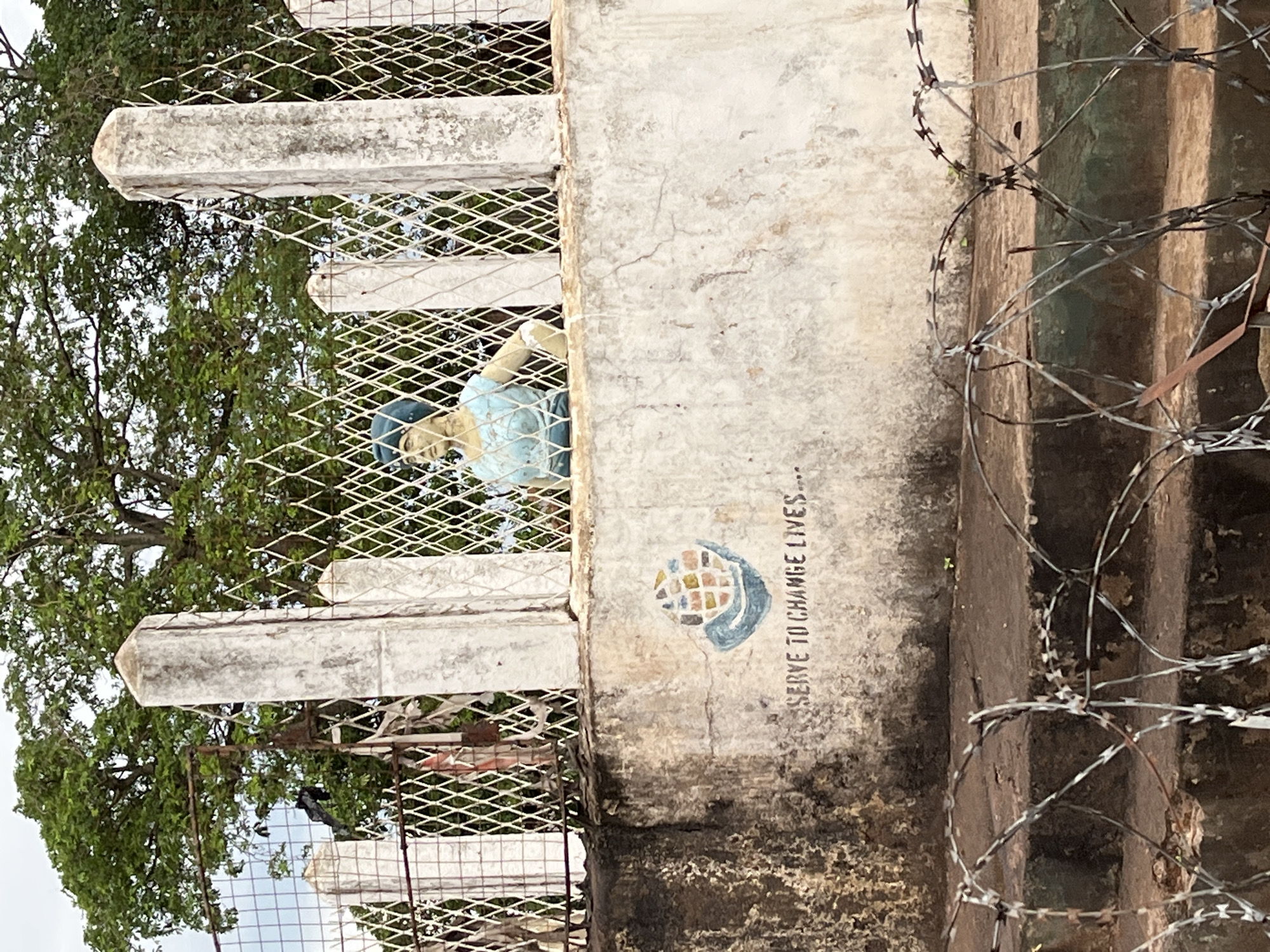 The image captures an outdoor scene in Idah, Nigeria, featuring a perimeter barrier and a statue. In the immediate foreground, coils of sharp razor wire are spread across a dark, multi-tiered concrete surface, likely steps or a low platform. Behind this, a weathered concrete wall rises, stained and showing signs of age. On the face of this wall, a light blue stylized globe cradled by a blue hand is painted, accompanied by the text "SERVE TO CHANGE LIVES..." in dark lettering. Above this wall, a security fence is constructed with white, diamond-patterned mesh supported by several rectangular concrete posts that are also weathered and discolored. Behind the fence and resting on the wall, a waist-up statue or mannequin of a person, likely female, is positioned. The figure wears a light blue sleeveless top and a dark blue head covering, looking forward with one hand on its hip. In the background, dense green foliage from large trees is visible, extending upwards towards a bright, overcast sky.