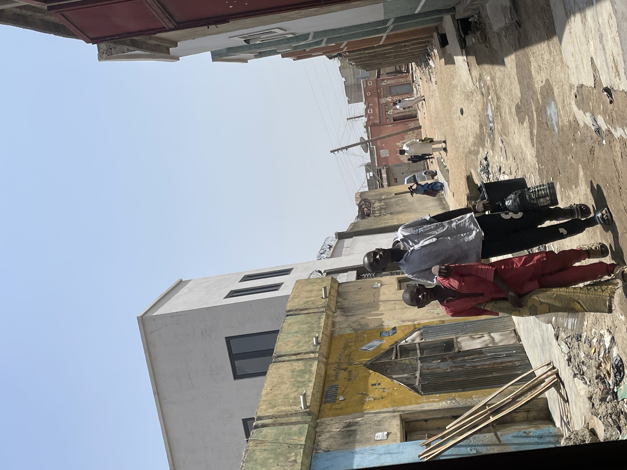 A street scene in what appears to be a lower-income urban area. Two people are positioned in the foreground. One is dressed in a light-colored shirt and dark pants and is carrying what appears to be a plastic container. The second person, possibly a child, is wearing red pants and a patterned shirt. The street is narrow, and there is visible litter and debris on the ground. Buildings flank both sides of the street, showing signs of age and varying architectural styles. In the distance, other people can be seen walking or standing. Overhead wires are present. The sky is light blue and clear.
