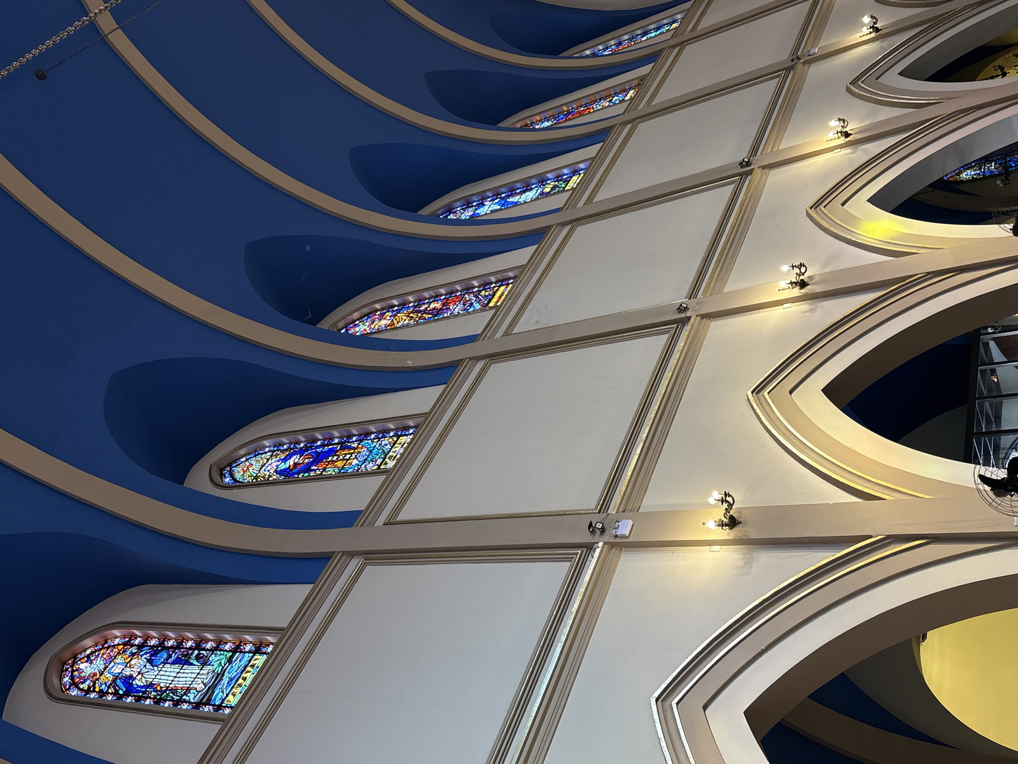 The image captures an interior view of a building, likely a church, featuring a high, arched ceiling painted a deep blue with cream-colored trim. Along the walls, several tall, narrow stained-glass windows are visible, depicting colorful religious scenes. The lower portion of the walls is adorned with a beige finish and framed by cream-colored arches. Wall-mounted light fixtures with multiple bulbs cast a warm glow. A fan is partially visible at the bottom of the frame. The architectural style suggests a grand, possibly historic, religious structure. Based on visual cues such as the architectural style and the prevalence of stained-glass windows, this scene is consistent with the interior of the Metropolitan Cathedral of São Paulo, Brazil.