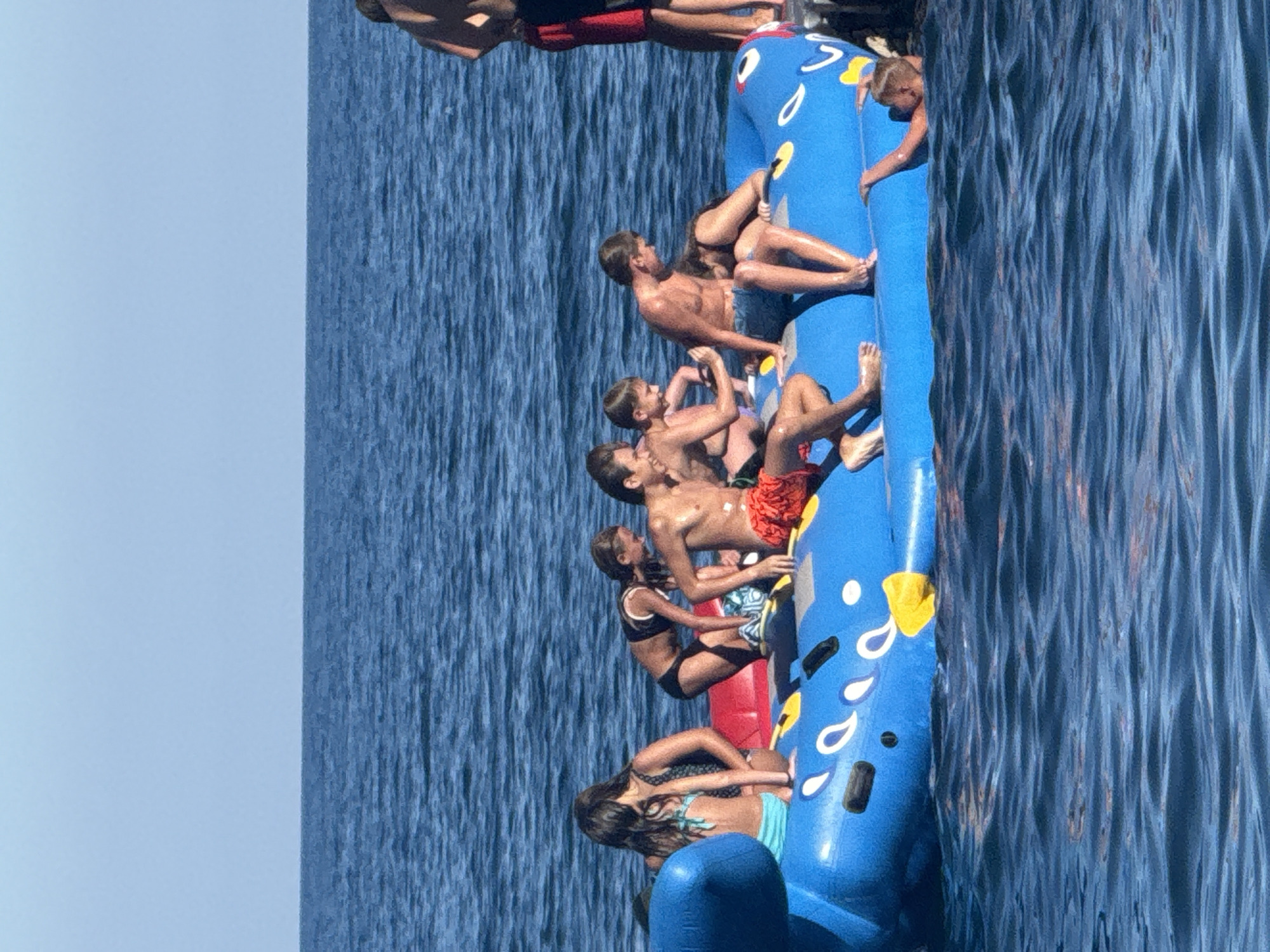 The image shows a group of people on a blue inflatable raft in the Black Sea. The raft has white and yellow decorations that resemble fish eyes and fins. Several individuals, appearing to be mostly teenagers and young adults, are seated or reclining on the raft. Some are wearing swimwear. The water is calm and blue, with a clear sky in the background. One person is partially submerged in the water to the right of the raft. The location is the sea off the coast of Partenit, Ukraine, a popular resort town.