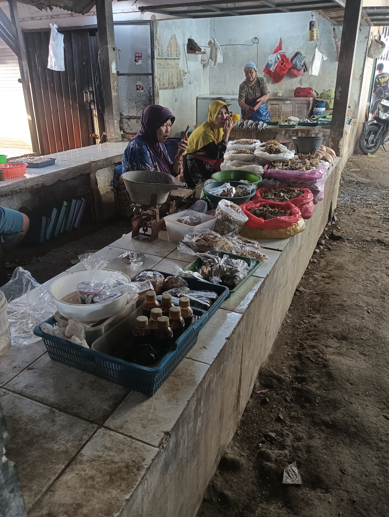 A street market stall in Cluwak, Indonesia, displays various dried goods. Three women are present. In the foreground, two women sit behind a table laden with bags of dried products, likely fish or seafood. One woman, wearing a purple hijab and blue patterned top, looks at her phone. The other, in a yellow hijab and black dress, rests her chin on her hand. Further back, a third woman, wearing a blue headscarf and patterned top, stands behind the counter, appearing to interact with something on the table. The stall features a scale, baskets filled with goods, and bottles of liquid. The environment is rustic, with a dirt floor and an open-air structure with a corrugated metal roof. A motorcycle is visible parked to the right of the stall.