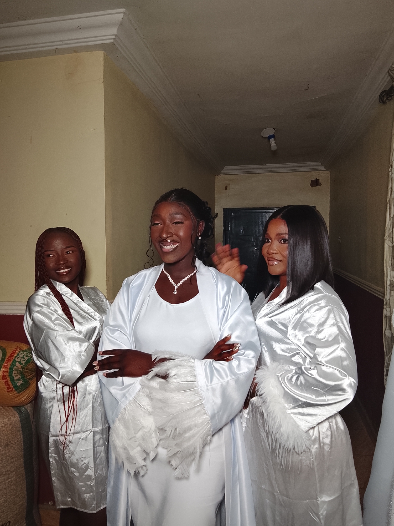 Three young Black women stand indoors. The woman on the left has braided hair and is wearing a white satin robe. She is standing with her arms crossed and smiling. The woman in the center is wearing a white dress with a white satin robe with feathers on the sleeves. She is smiling and has her arms crossed. The woman on the right is wearing a white satin robe and has shoulder-length black hair. She is smiling and raising her hand. The women are in a hallway with beige walls. There is a light fixture on the ceiling. The location is Unknown, Unknown.