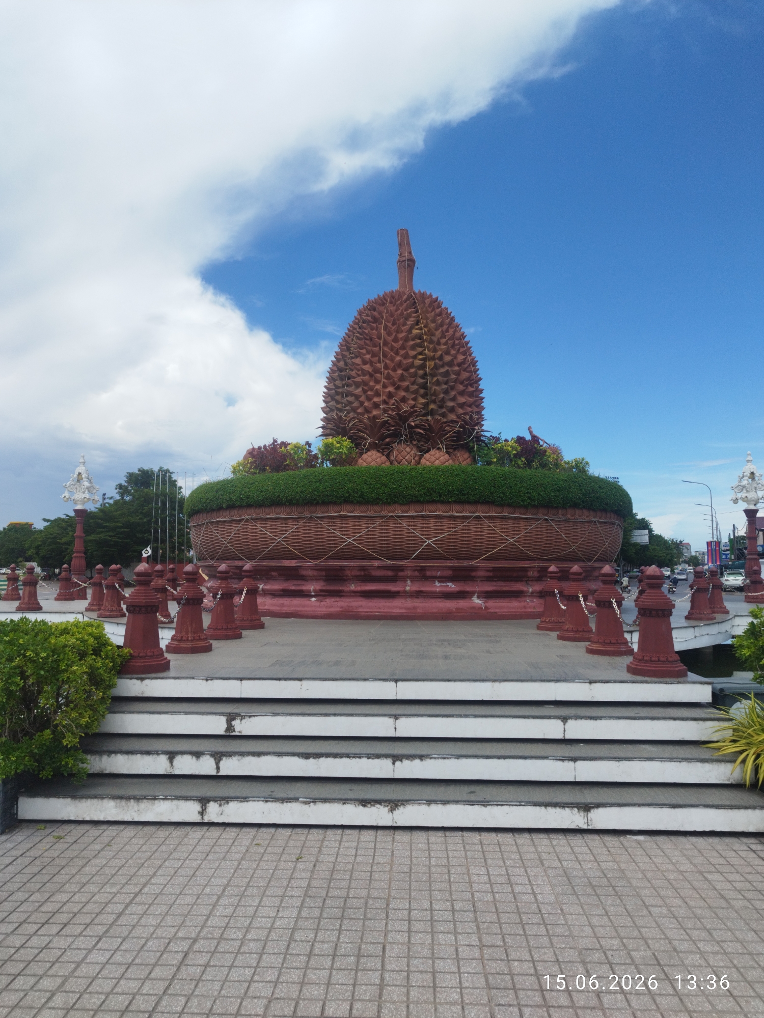 The image depicts the Durian Roundabout monument in Kampot, Cambodia. The monument, shaped like a large durian fruit, sits atop a circular base resembling a woven basket. It is surrounded by red bollards with chains and ornate white lampposts on either side. Green shrubs and foliage are planted around the base, and a set of white-bordered stairs leads up to the monument. The sky is blue with scattered white clouds. In the background, trees, buildings, streetlights, and traffic can be seen, indicating an urban environment. The timestamp "15.06.2026 13:36" is visible in the lower right corner. There are no people visible in the image.
