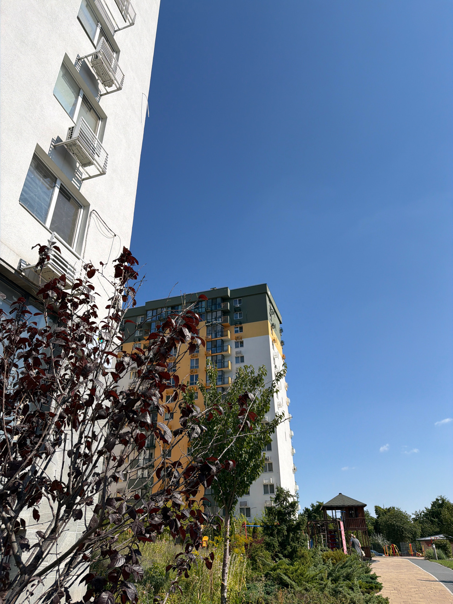 The image captures a scene in a residential area of Kyiv, Ukraine, on a clear day with a bright blue sky. In the foreground, a modern white apartment building stands tall, its facade punctuated by numerous windows and air conditioning units mounted on individual balconies. To the right of this building, another taller apartment building is visible, characterized by its colorful exterior featuring yellow, white, and green sections. A prominent element in the middle ground is a children's playground with a wooden playhouse and various structures. Lush green trees and bushes surround the playground and extend towards the foreground, where a tree with dark reddish-brown leaves is prominently featured. The ground in the foreground consists of a paved walkway with a brick-like pattern, suggesting a well-maintained urban outdoor space. The overall atmosphere is one of a bright, contemporary urban environment with recreational facilities.