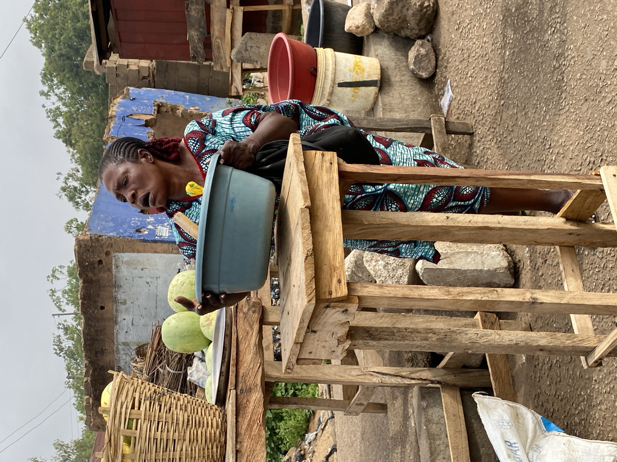 In Bukuru, Nigeria, a woman sits on a crude wooden structure, possibly a makeshift stool or display. She wears a patterned, colorful dress and her hair is styled in braids or twists. She holds a light blue basin or container, possibly used for washing or displaying goods. Beside her are several green fruits, likely watermelons, arranged on a surface. A woven basket filled with yellow fruits is also present. The background shows a partially dilapidated building with blue paint. The environment appears to be an outdoor market or roadside selling area, with a mix of natural and constructed elements. Other items include buckets, stones, and a sack.