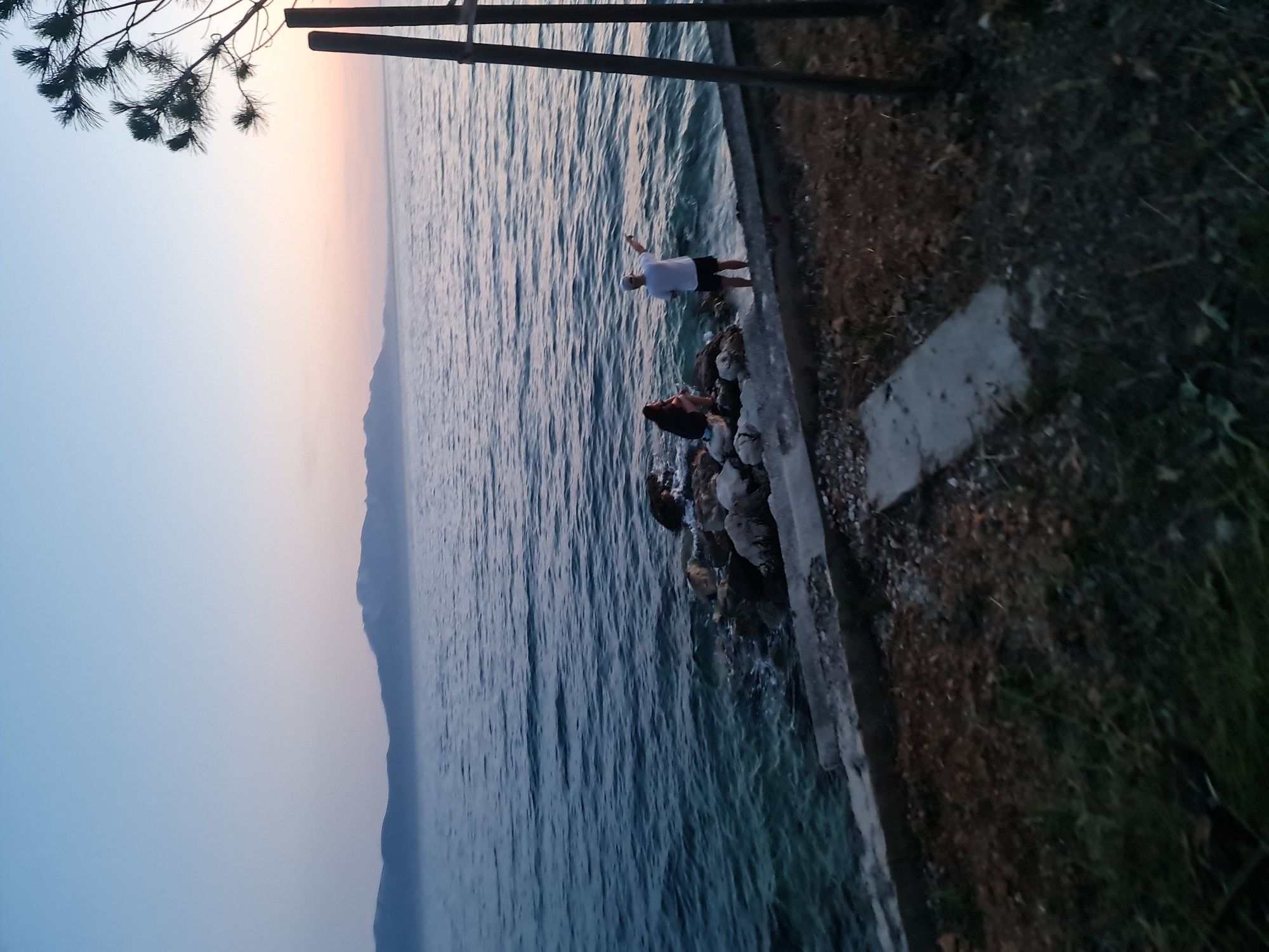 The image depicts a serene twilight scene by the sea in Brist, Croatia. The sky transitions from a pale blue at the top to soft hues of pink and orange near the horizon, with distant mountains silhouetted against the fading light. The calm sea stretches out, reflecting the subtle colors of the sky.
In the foreground, a rocky shoreline meets a concrete walkway. Two individuals are present near the water. A person in a dark top and shorts is seated on the rocks, while another person in a white t-shirt and dark shorts stands at the water's edge, gesturing towards the sea. To the right, the dark silhouettes of two wooden posts stand against the twilight sky, with a few branches of a pine tree visible at the top right corner. The ground in the immediate foreground consists of dry grass and scattered stones.