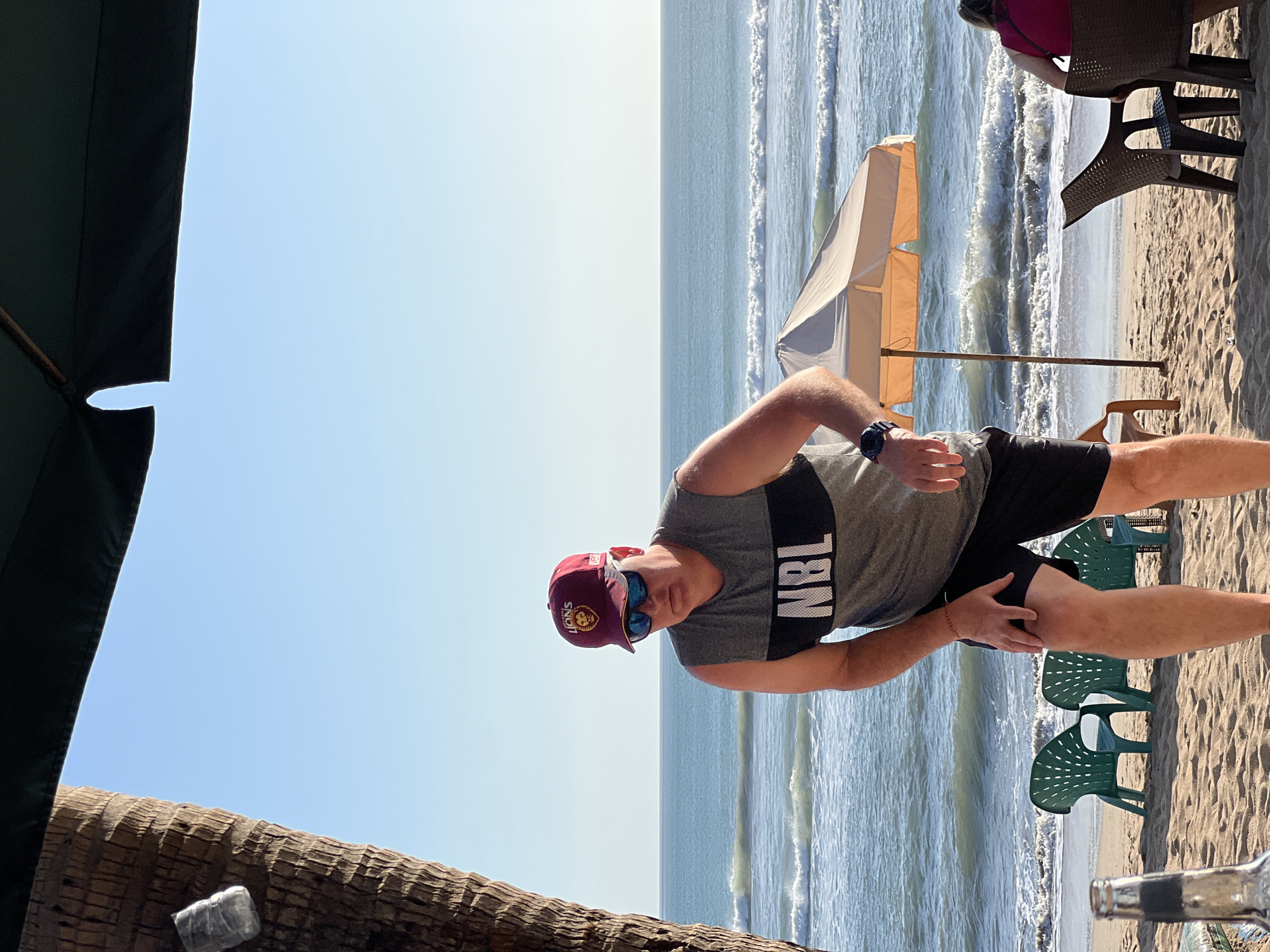 A man wearing a maroon baseball cap with a lion logo, sunglasses, a gray sleeveless t-shirt with "NBL" printed in large white letters on the chest, and black shorts is standing on a sandy beach. He is looking down and slightly to his left, with his right hand resting on his right knee. He appears to be in motion, possibly bending down.
The beach overlooks the ocean with moderate waves. In the background, there are two green plastic chairs and a closed light-colored beach umbrella. Further in the distance, two people are seated at chairs facing the ocean. A palm tree trunk is visible on the left side of the frame, and a partial view of a dark green umbrella is above. The sky is clear and blue.
This scene is characteristic of the beaches in Kuta, Bali, Indonesia, where such beachside setups with umbrellas and chairs are common for tourists and locals enjoying the ocean.