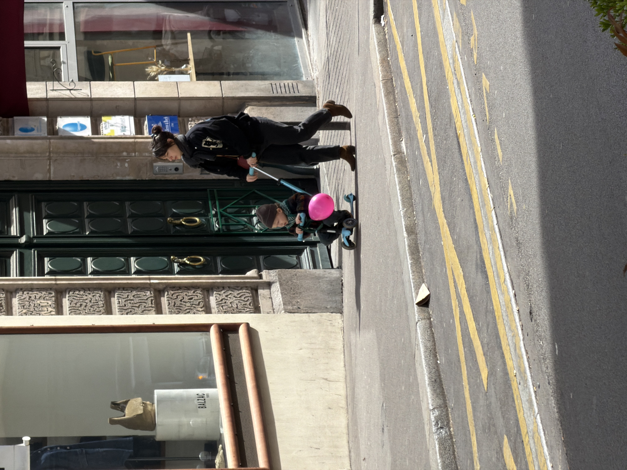 A mother pushes her child on a scooter along a street in Paris, France. The child sits on the scooter with a pink balloon attached to the handlebars. The street is marked with yellow lines. The architectural style suggests a Parisian street. A storefront with a visible “Balzac” label on a canister in the display window indicates a local business.