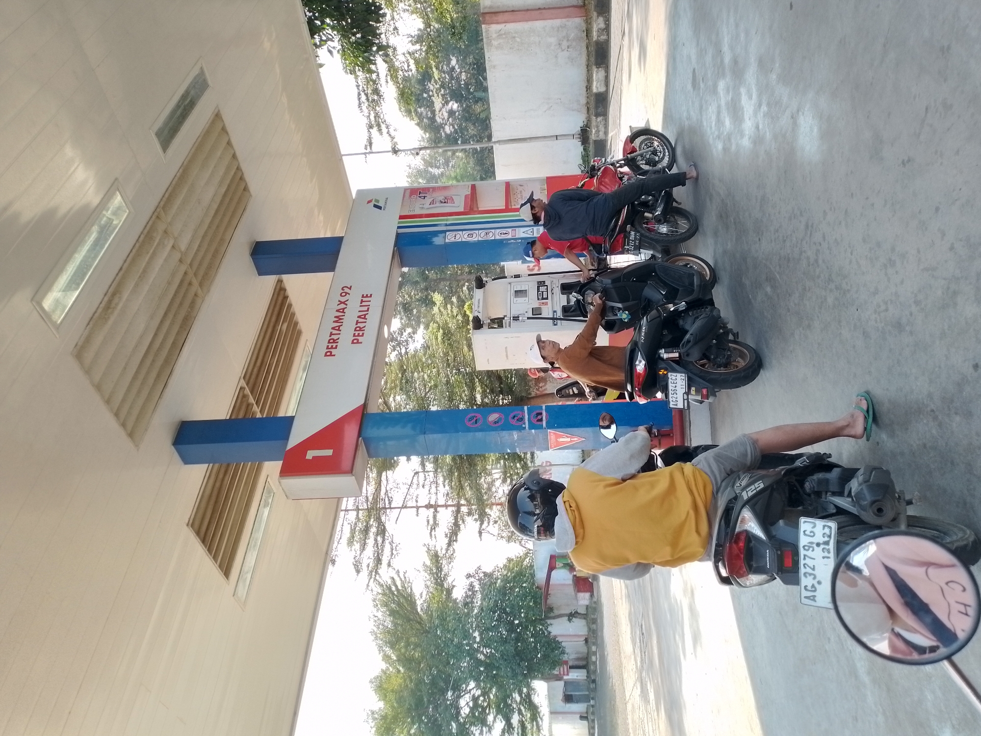 The image depicts a scene at a Pertamina gas station in Ngancar, Indonesia. A group of individuals on motorcycles are waiting in line or are in the process of refueling. In the foreground, a person wearing a yellow hoodie and a helmet is seated on a black motorcycle. The motorcycle has a license plate that reads "AG 3279 GJ 12-27". To the right of this motorcycle, another black motorcycle is parked, and a person in a brown shirt is standing beside it. Behind them, a red motorcycle is visible with a person seated on it. A gas pump with the Pertamina logo and information about "PERTAMAX 92 PERTALITE" is present in the background, along with a blue and white support pillar. The environment appears to be an outdoor gas station with a covered roof, and trees are visible in the background under a bright, clear sky. The ground is paved with concrete.