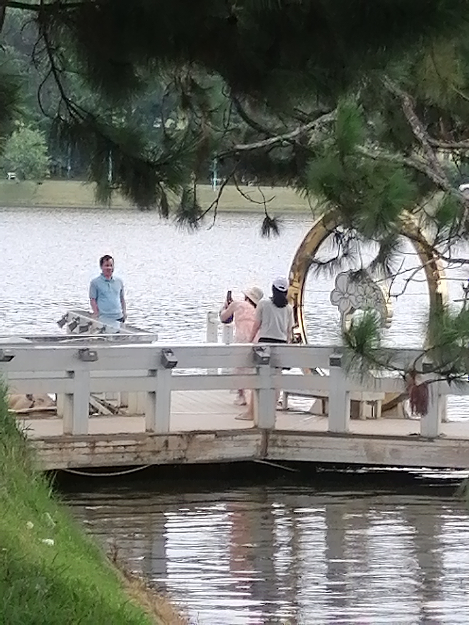 The image depicts a scene at Xuan Huong Lake in Da Lat, Vietnam. A wooden pier extends into the water, with several people on it. On the left side of the pier, a man in a light blue polo shirt and white pants stands and smiles at the camera. In the center of the pier, a woman in a pink shirt and white hat is taking a photo. To her right, another woman in a white shirt and a dark cap is looking away. Behind them, a decorative archway with a flower motif is visible over the water. The water is rippling, reflecting the sky and surrounding trees. The foreground shows a grassy embankment on the left. The overall atmosphere suggests a casual outing at a popular tourist spot.
