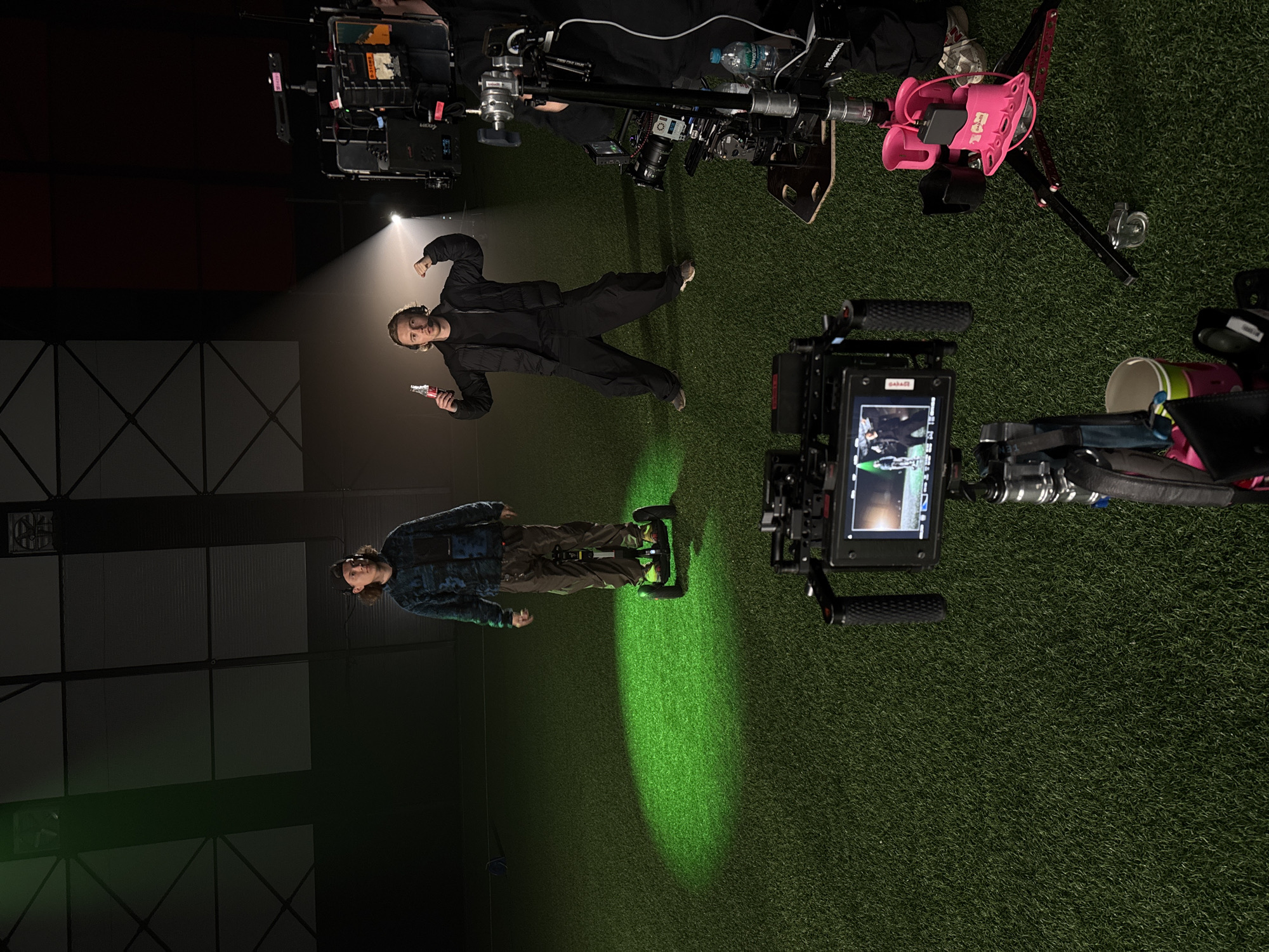 The image captures a behind-the-scenes moment on what appears to be a film or video set. The scene is likely indoors, possibly in a studio or large space with a grid-like ceiling structure visible.
Two figures are prominently featured:
* One individual is wearing a black outfit and holding a small, red object (possibly a toy gun). They are standing under a bright, focused spotlight.
* Another individual is wearing a blue and green plaid shirt and brown pants and standing on a dolly. They are illuminated by a green spotlight.
The floor is covered in what looks like artificial turf. The image also includes film equipment such as cameras, tripods, and other supporting gear. A pink accessory or part is mounted on a tripod.
There is no direct evidence in the image to confirm the location is Balashikha, Russia. The description is based solely on the content of the image.