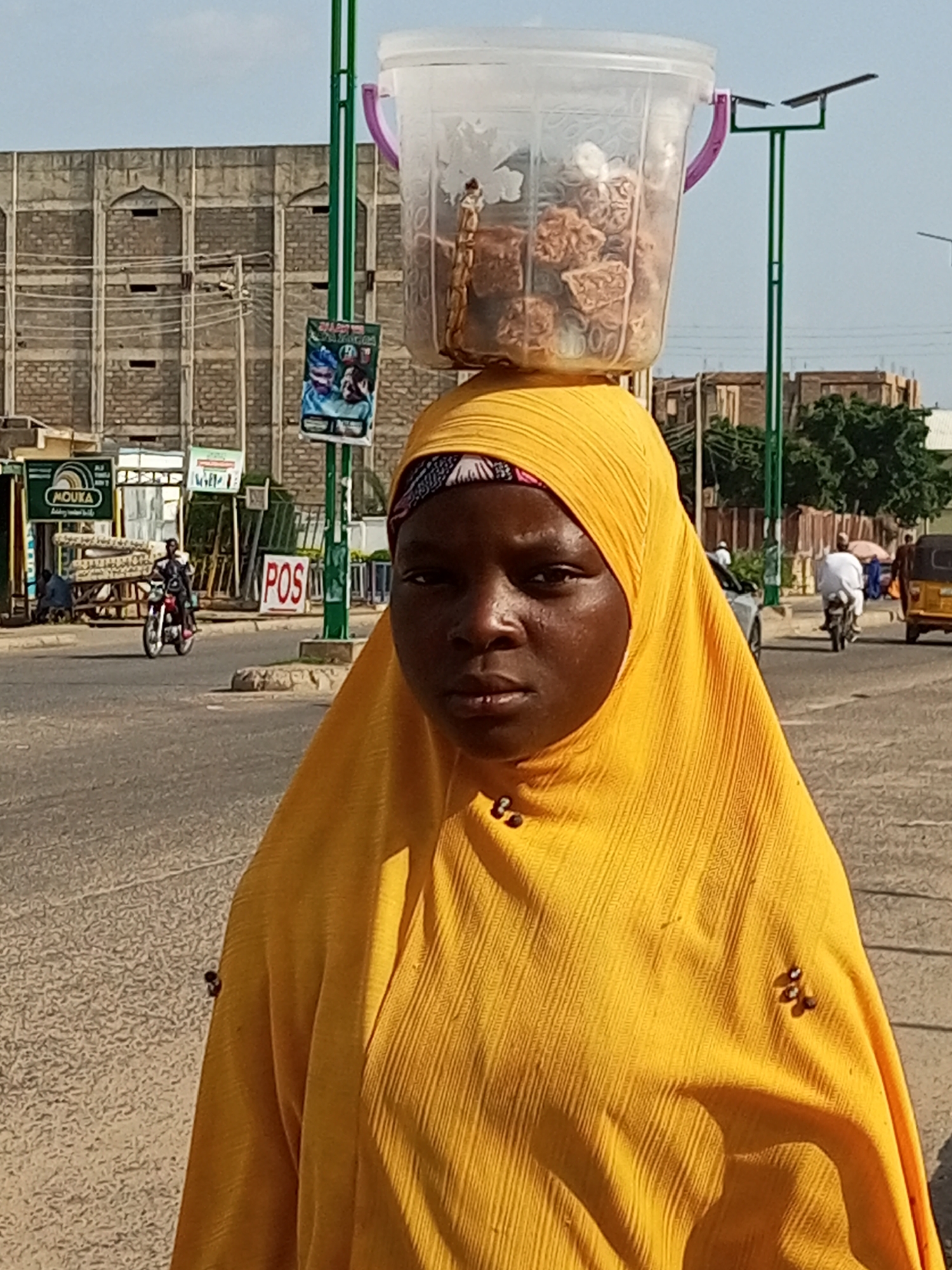 In Arawa, Nigeria, a young woman wearing a bright yellow headscarf and matching garment, stands in the foreground of a street scene. She carries a transparent plastic bucket filled with what appears to be food items, balanced on her head. The bucket has a pink handle and is partially obscured by her headscarf. Behind her, a paved road stretches into the distance, with various vehicles and pedestrians visible. To the left, a building with distinctive arched windows and brickwork suggests an urban setting. A green lamppost with a solar panel is positioned to the right of the woman. The overall atmosphere is that of a busy marketplace or a street in a Nigerian city.