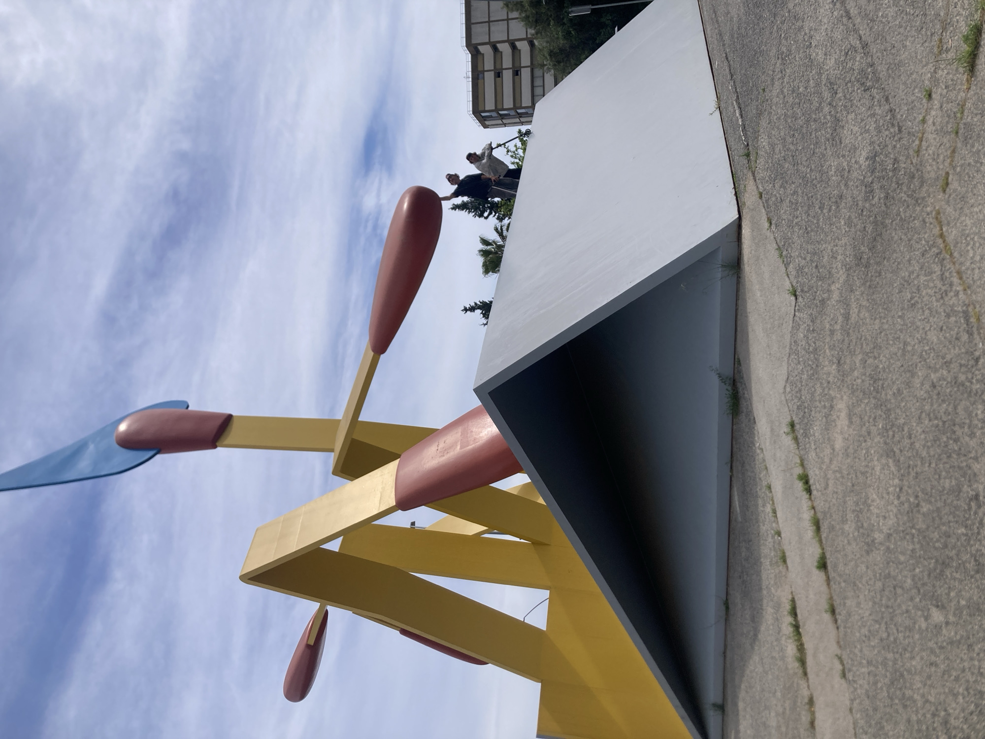 The image shows the sculpture "Els Mistos" (The Matchsticks) in Barcelona, Spain. The sculpture is made of yellow, red, and blue painted metal. The image is taken from a low angle, looking up at the sculpture against a sky with light clouds. In the background, there is a white, box-like structure with an opening, and a building beyond it. Two people are standing on a ledge near the building, appearing to be interacting with the sculpture. The ground is concrete with grass growing in cracks.
