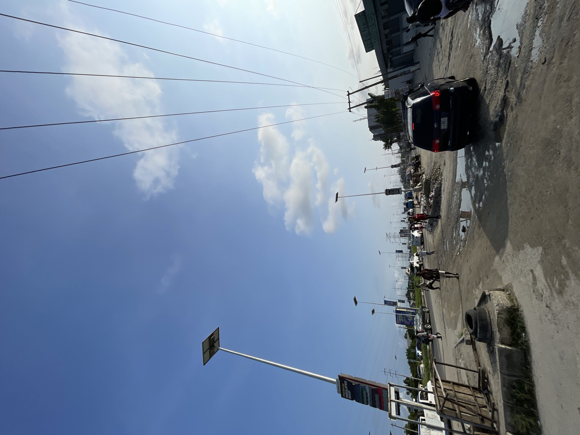 This is a ground-level, eye-level shot of a street in Lagos, Nigeria. The sky is bright blue with scattered white clouds. Overhead, multiple power lines stretch across the frame, supported by utility poles in the distance. On the left side of the street, a tall, modern solar-powered streetlamp stands prominently, adorned with a billboard advertising "PREMIUM INSULATED ROOF." Several other similar streetlamps are visible further down the road.
The street itself is unpaved and appears to be in poor condition, with numerous large puddles and potholes evident. A black SUV is driving through the puddles, its taillights illuminated. Several people are walking along the side of the road, some appearing to be engaged in conversation or going about their daily activities. On the right side of the street, various buildings line the road, some with visible signage. The overall impression is of a busy, developing urban environment with ongoing infrastructure challenges.