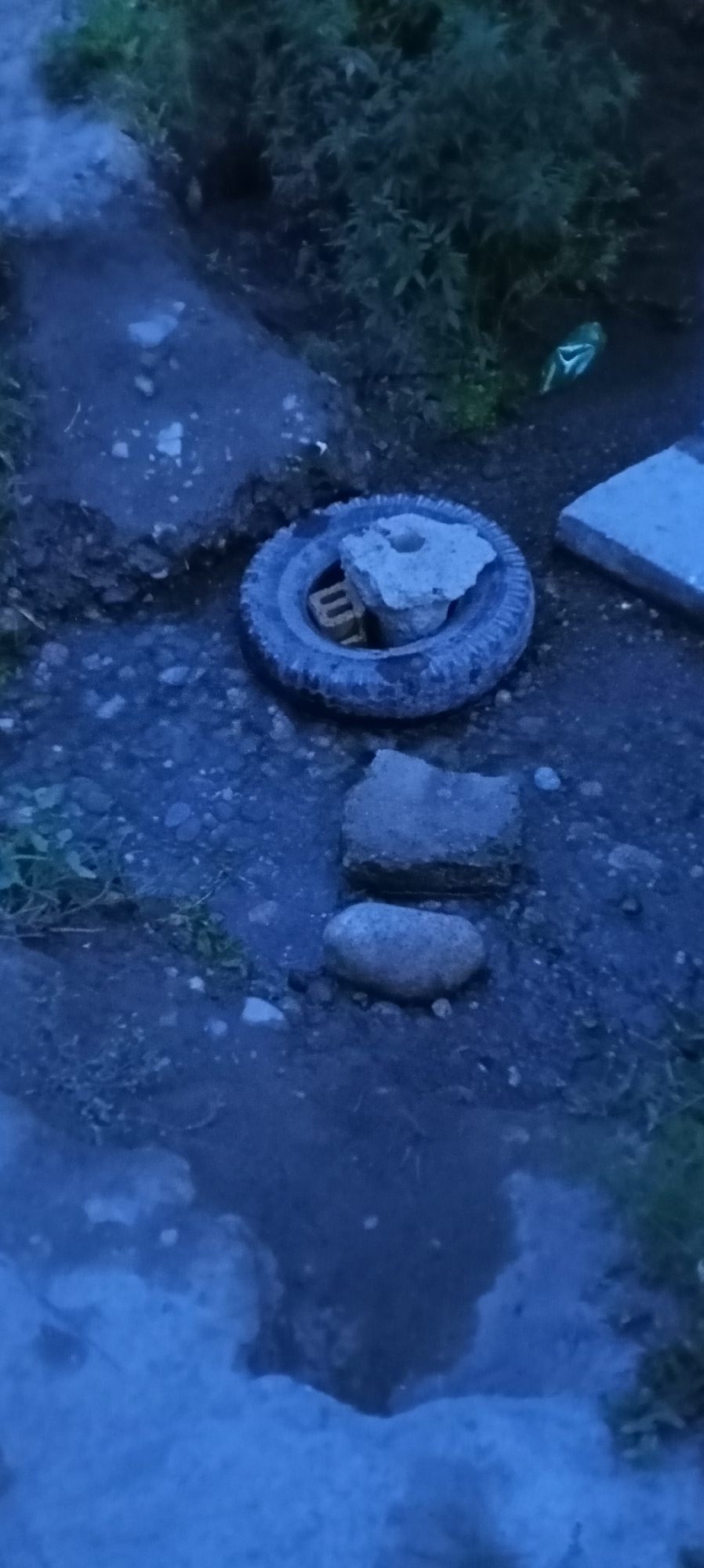 The image shows an outdoor scene with a tire lying flat on the ground. A piece of concrete is resting on the tire, and part of a brick is visible inside the tire, underneath the concrete. Two flat stones are also on the ground near the tire. The ground appears to be a mixture of dirt, gravel, and some sparse vegetation. In the background, there are bushes and foliage. A blueish tint to the image suggests it was taken at dusk or in low light conditions. The scene is consistent with an informal outdoor area, possibly a construction or work site, or a yard in Sokuluk, Kyrgyzstan.
