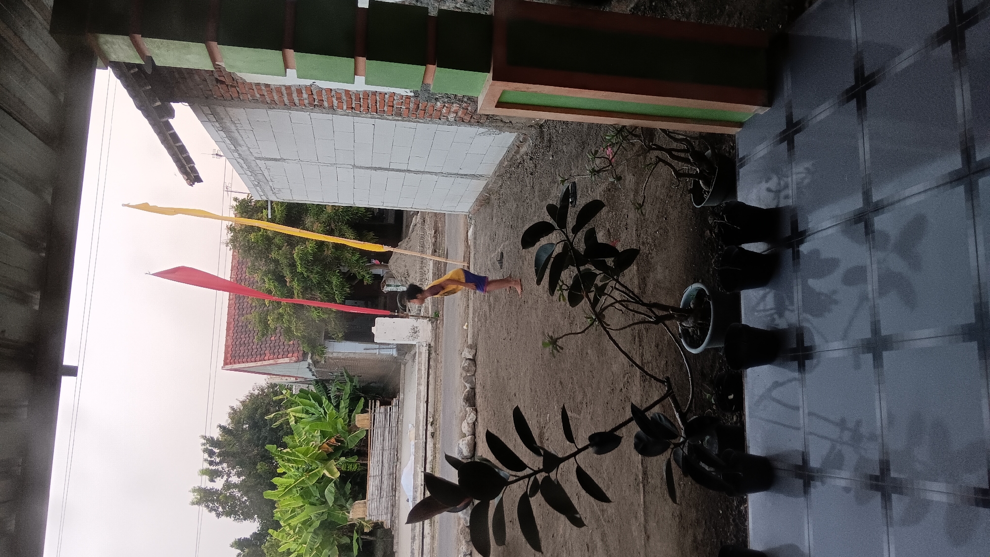 In the city of Sonobekel, Indonesia, a young boy wearing a yellow shirt and blue shorts stands barefoot on a dirt ground. He is facing away from the camera and appears to be looking at something off-frame to his right. Two flags, one red and one yellow, are prominently displayed behind him, tied to poles. In the foreground, several potted plants are arranged on a tiled patio. The scene is framed by a roofed structure with supporting pillars, one of which is decorated with green and orange accents. The background shows residential buildings, trees, and a street. The sky is overcast.