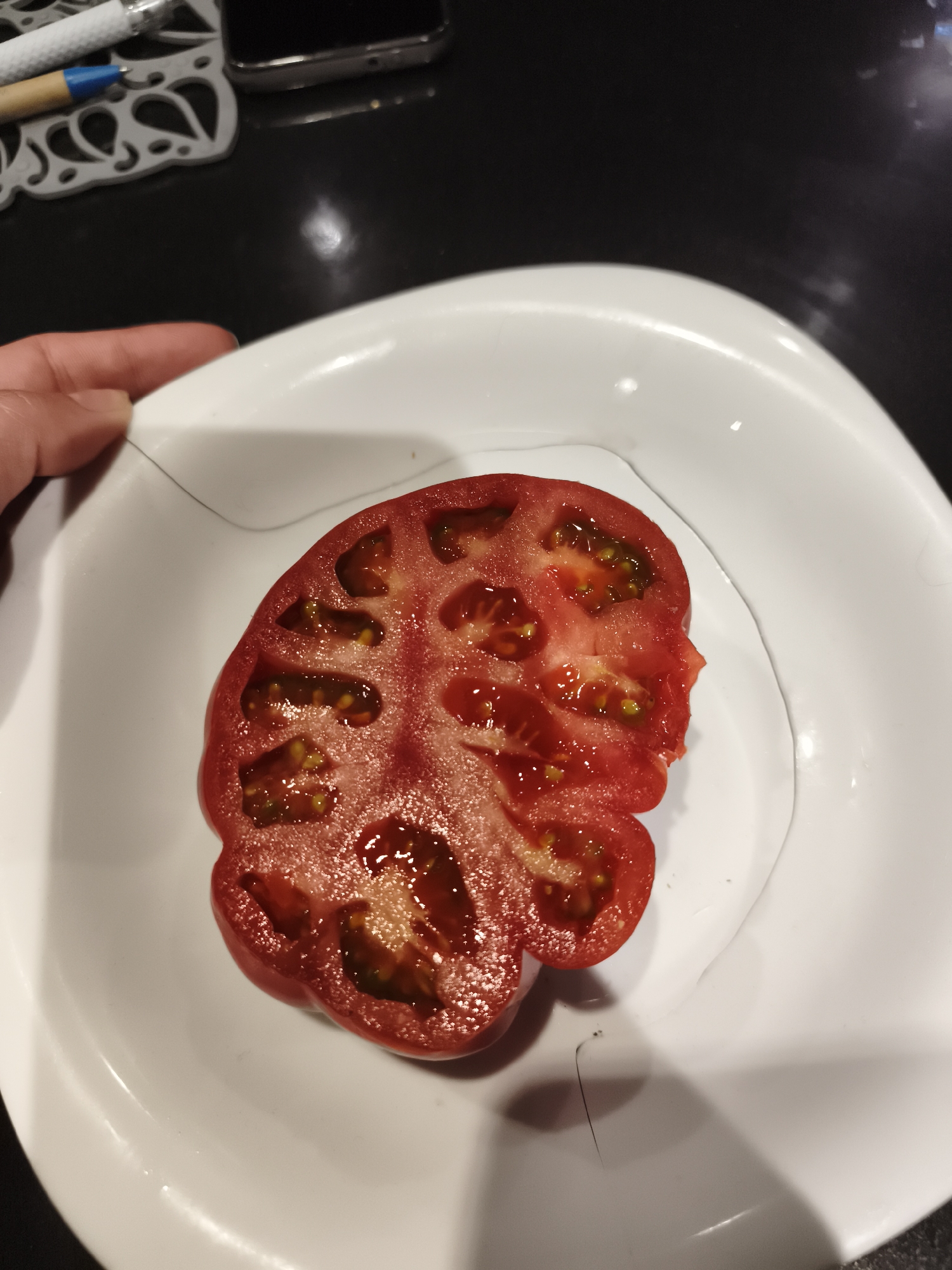 A close-up, overhead shot depicts a vibrant red tomato, sliced in half horizontally, resting on a white, irregularly shaped plate. A hand partially visible on the left holds the plate. The tomato's cross-section reveals numerous internal chambers (locules) filled with seeds and translucent gel, characteristic of a juicy beefsteak or heirloom variety. Its form is organic and slightly lobed.
The white plate sits on a dark, glossy countertop or table surface. In the blurred background, a smartphone, a white pen with a blue tip, and a decorative white coaster are faintly visible, suggesting an indoor setting, likely a kitchen.
Notably, two distinct strands of dark hair are present on the white plate; one long, thin strand stretches from near the holding hand across the upper left portion of the plate, and a shorter, curlier piece lies in the bottom right corner. The lighting appears to be artificial, casting soft reflections on the wet tomato surface and the glossy countertop. There is no visible text in the image. The scene captures a moment of food preparation in what appears to be a domestic setting in Burgas, Bulgaria.
