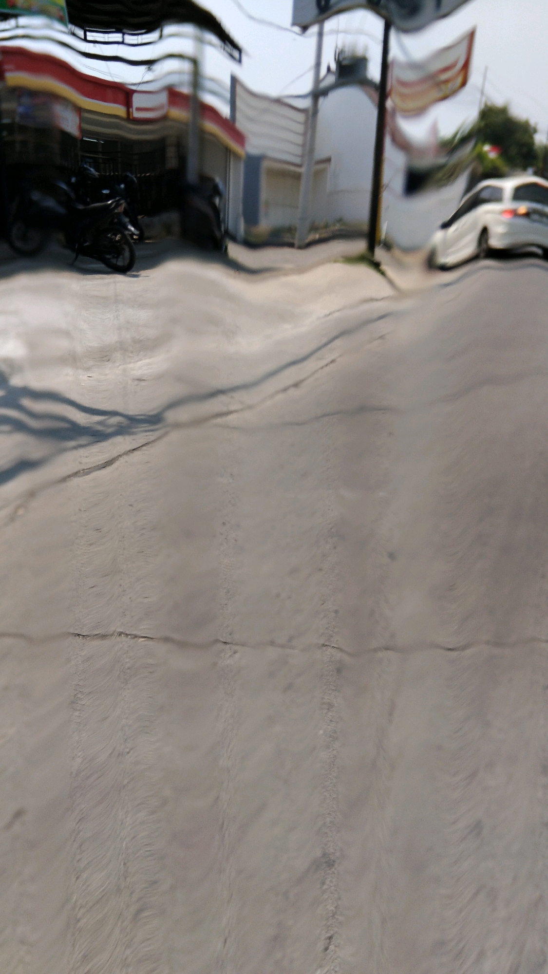 The image shows a street scene in Watesalit, Indonesia, with a slight visual distortion that warps the horizontal lines of the buildings and road. In the foreground, a gray asphalt road with visible cracks and texture is depicted. Shadows of objects, likely from a nearby structure, are cast across the road. In the background, a row of buildings lines the street, one of which appears to be a storefront with a red and yellow awning. Two dark-colored motorcycles are parked in front of the storefront. Further down the street, a white car is visible. The sky is bright and appears to be daytime. The overall scene suggests a typical urban or suburban environment in Indonesia.